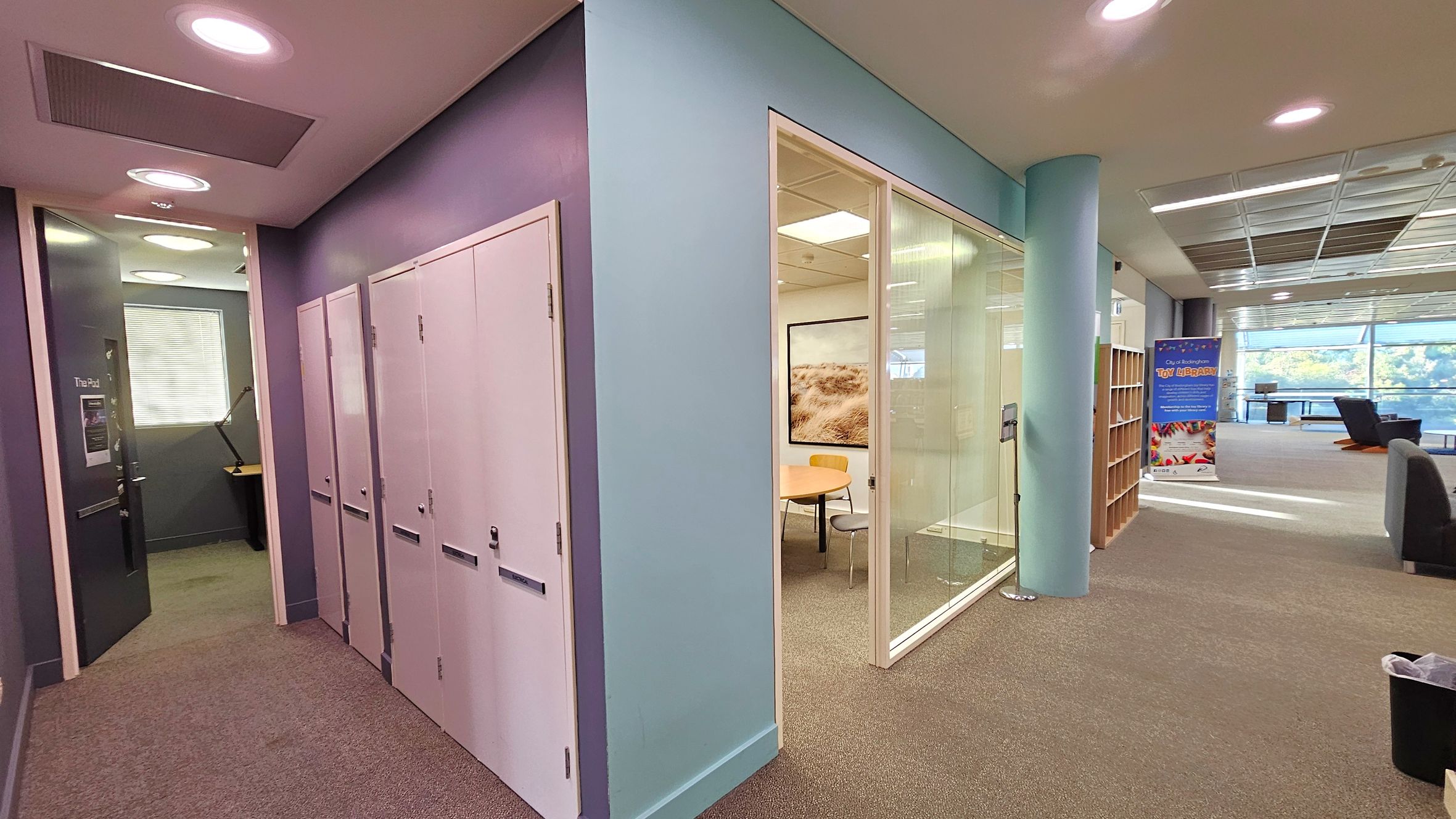 A modern office space with a corridor on the left featuring white lockers and a door labelled "The Pod" leading to a small room with a desk and lamp. In the centre, a glass-walled meeting room contains a round table and chairs. On the right, an open area with bookshelves, a colourful banner, and seating near large windows. The ceiling has recessed lighting, and the floor is carpeted.