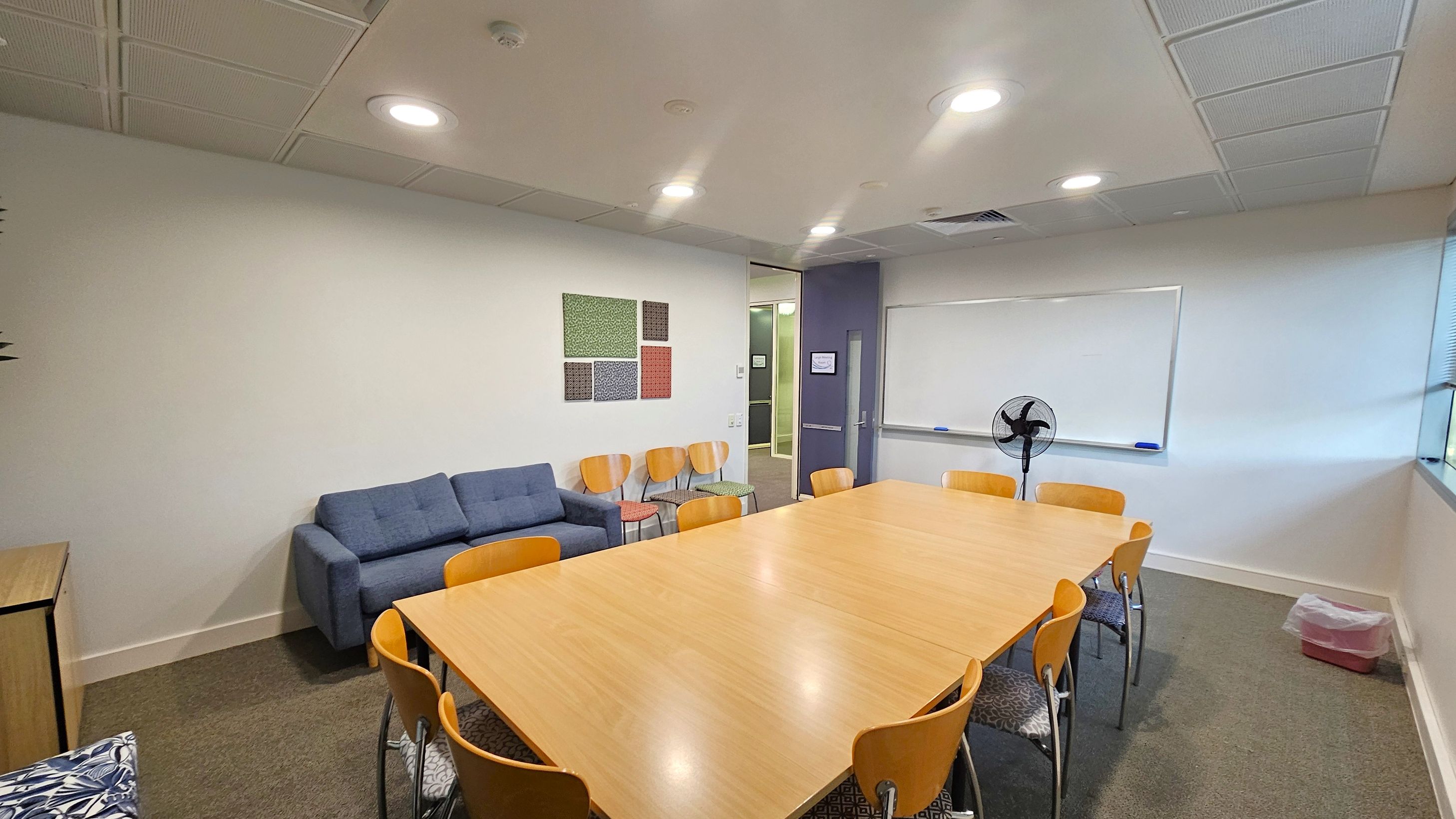 A meeting room with a large wooden table surrounded by wooden chairs in the centre. On the left, there's a blue sofa and several wooden chairs against the wall, with decorative panels above. On the right, a whiteboard is mounted on the wall with a fan in front of it. A small bin is on the floor near the window. The ceiling has recessed lighting, and the floor is carpeted.