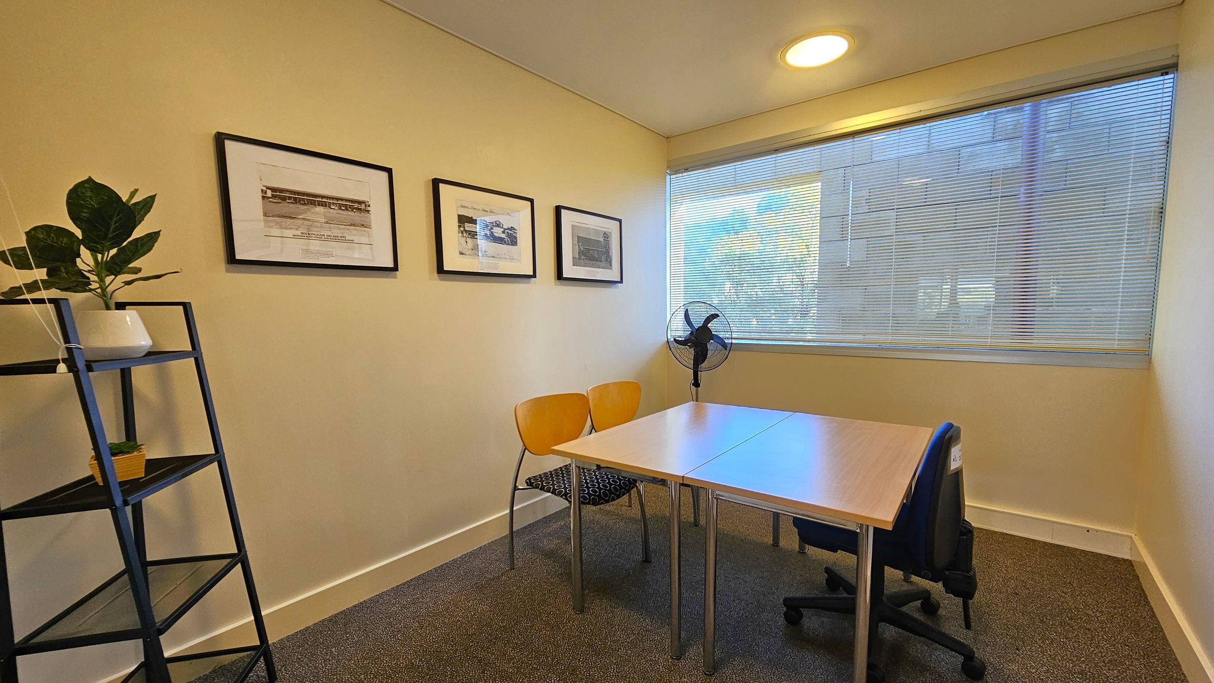 A small office room with a light-coloured carpeted floor. On the left, a black metal shelf holds two potted plants. The left wall displays three framed pictures. In the centre, a wooden table is surrounded by two yellow chairs and a blue office chair. On the right, a large window with blinds partially open allows natural light in. A black fan is positioned near the window. The ceiling is plain with a round light fixture.