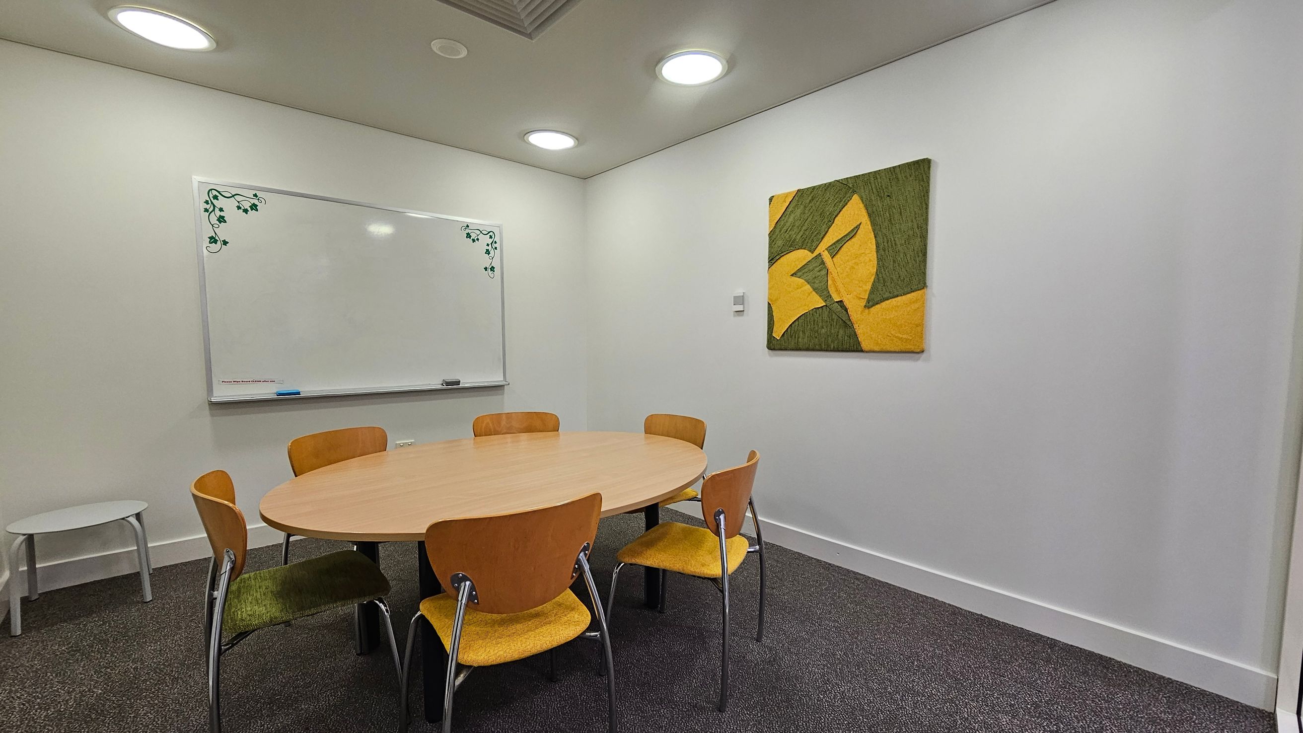 A small meeting room with a round wooden table surrounded by five wooden chairs with yellow seats. On the left wall, there's a whiteboard with green vine decorations in the corners. A small white side table is also on the left. The right wall features a green and yellow abstract painting. The floor is carpeted in dark grey, and the ceiling has recessed circular lights.