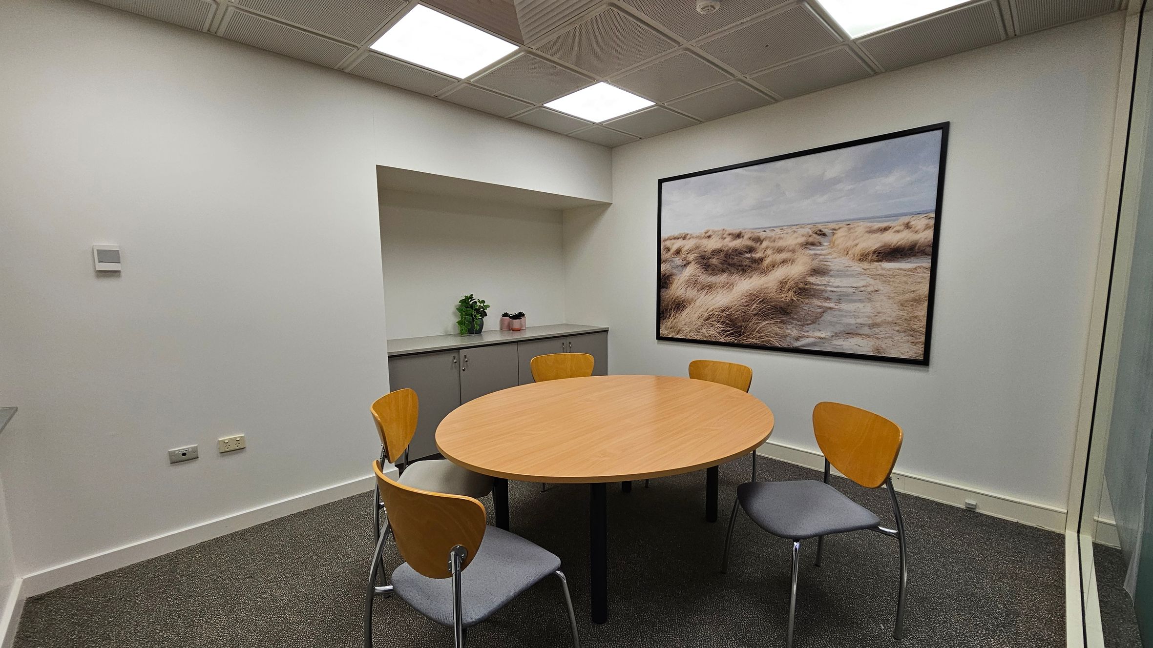 A small meeting room with a round wooden table surrounded by four chairs with wooden backs and grey seats. On the right wall, there's a large framed photograph of a grassy landscape. The back wall has a grey cabinet with two potted plants on top. The ceiling features recessed lighting panels, and the floor is carpeted in dark grey. The walls are painted white.