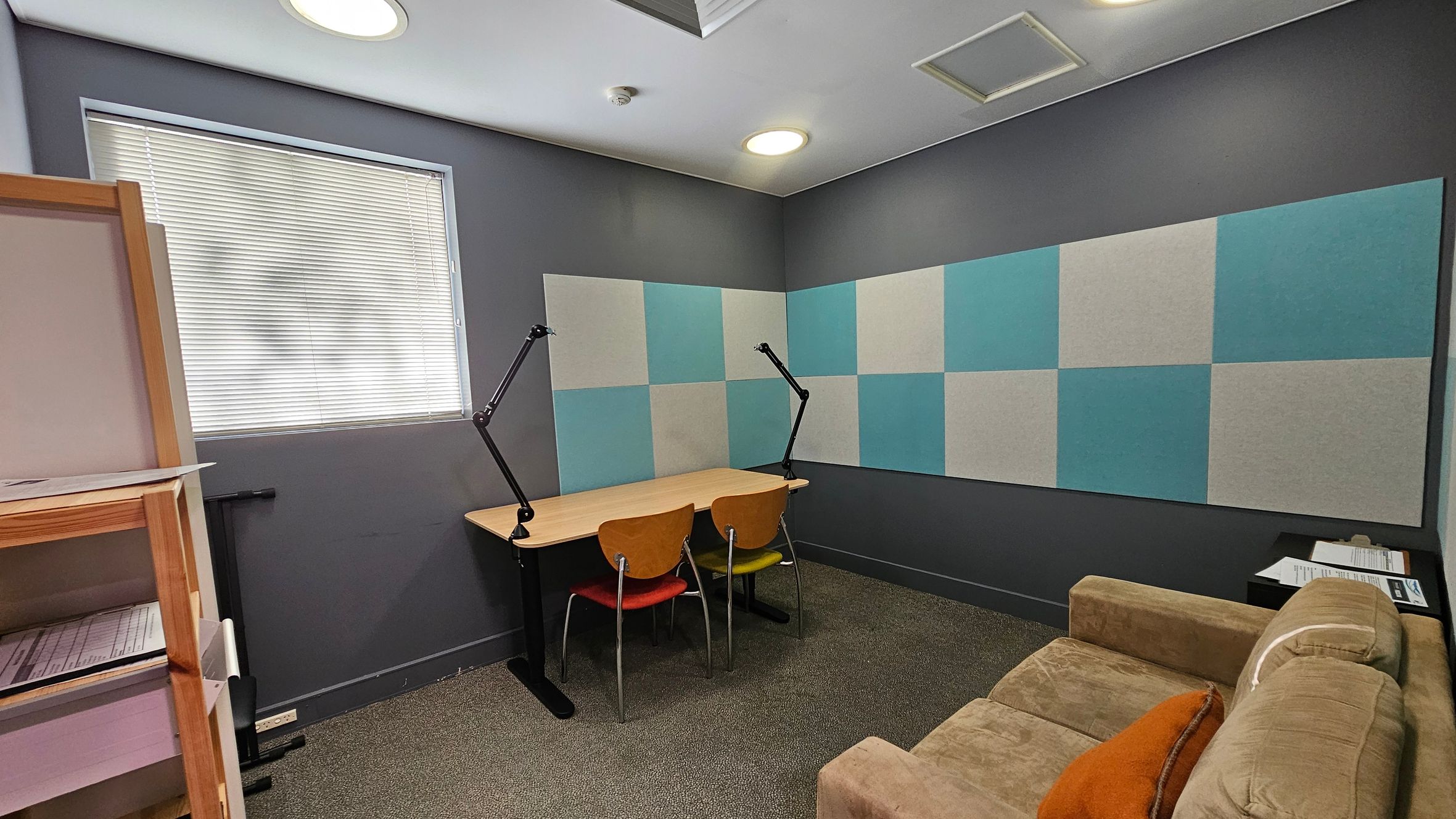 A small room with grey walls and a window with closed blinds on the left. Below the window is a wooden shelf with papers. In the centre, a wooden desk with two chairs and adjustable lamps. The wall behind the desk has a pattern of alternating blue and grey panels. On the right, a brown sofa with an orange cushion and a small black table with papers. The floor is carpeted in a mix of brown and green, and the ceiling has recessed lighting.