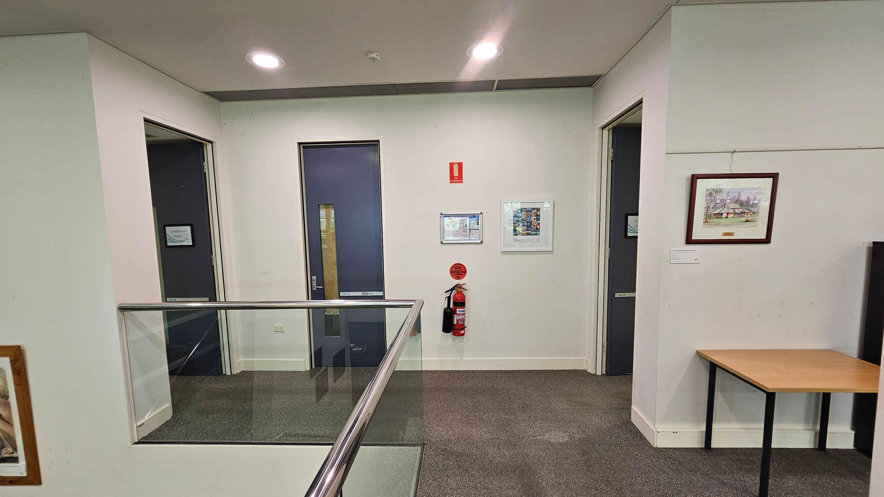 A hallway with a grey carpeted floor and a light-coloured ceiling with recessed lights. On the left, there is a glass railing and an open door labelled "Conference Room." In the centre, a closed dark door is flanked by a fire extinguisher and framed notices on the wall. On the right, another open door and a framed painting are visible above a small wooden table.
