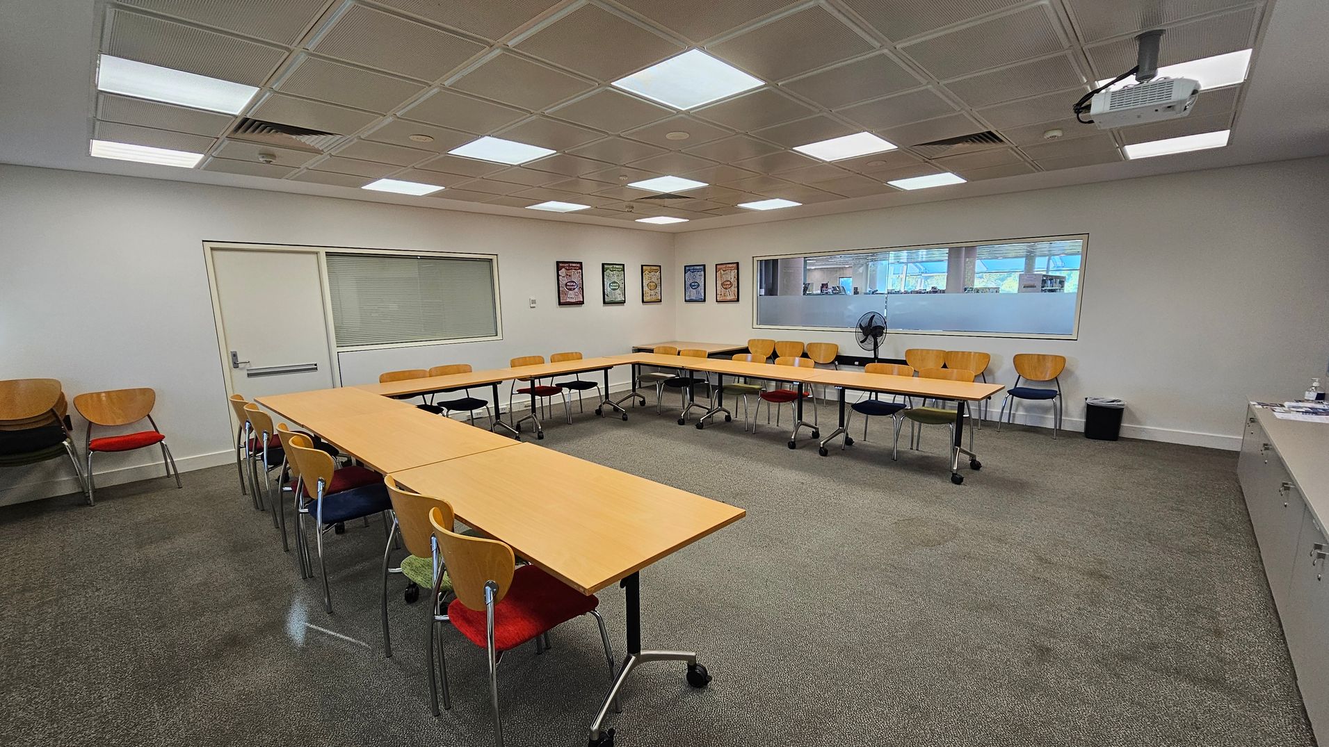 A conference room with a U-shaped arrangement of wooden tables and chairs. The floor is carpeted in green. On the left, there are stacked chairs with red seats. The back wall features framed posters and a large window with blinds. A fan is placed on a table against the wall. The ceiling has recessed lighting and a mounted projector. On the right, there is a long cabinet with various items on top.