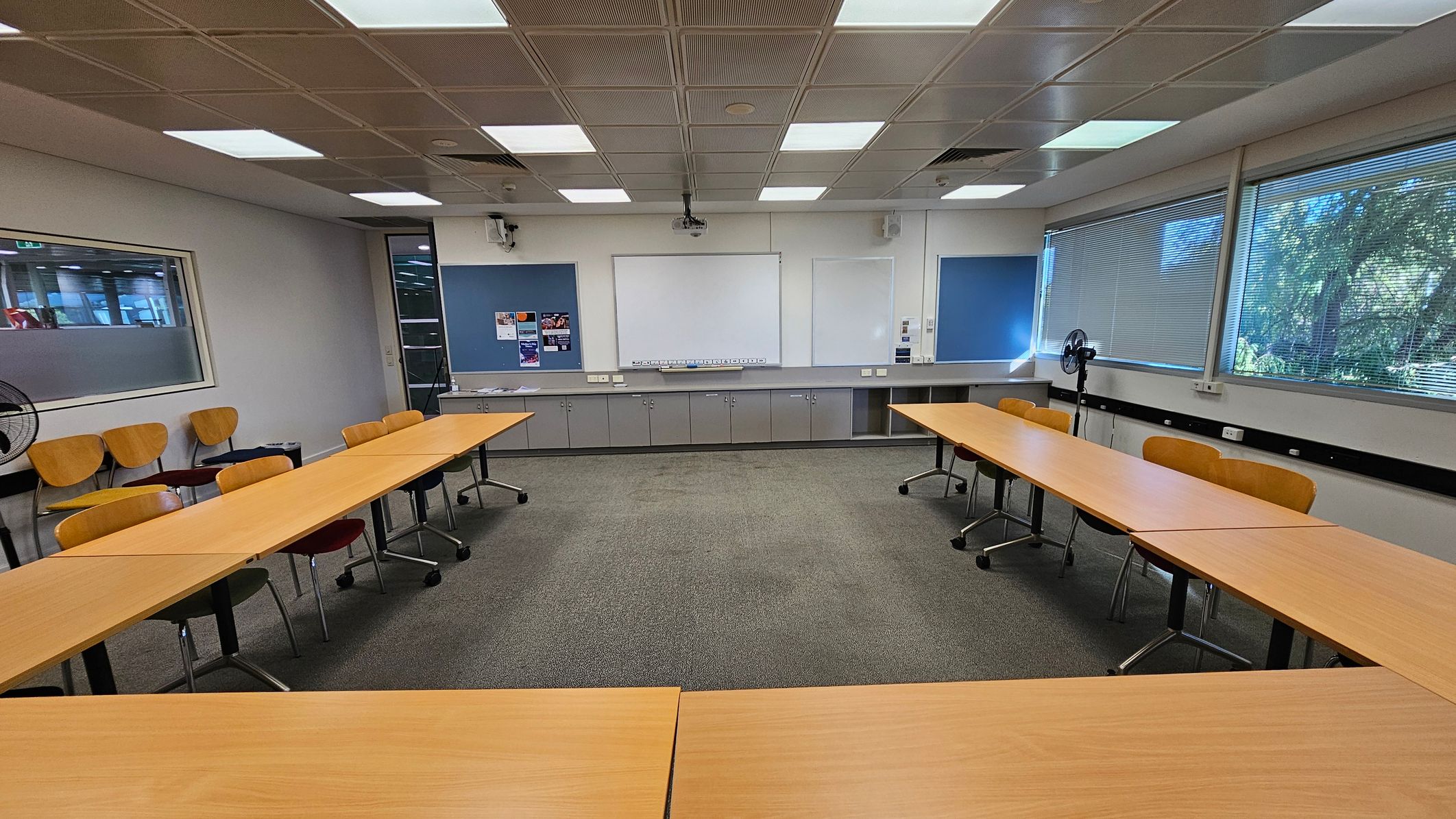A classroom with a U-shaped arrangement of wooden tables and chairs on a grey carpeted floor. The back wall features a large whiteboard flanked by blue noticeboards with some posters. Below the boards are grey cabinets. On the right, large windows with blinds allow natural light in, and a fan is positioned nearby. The ceiling has a grid pattern with fluorescent lights. On the left, a glass panel separates this room from another area.