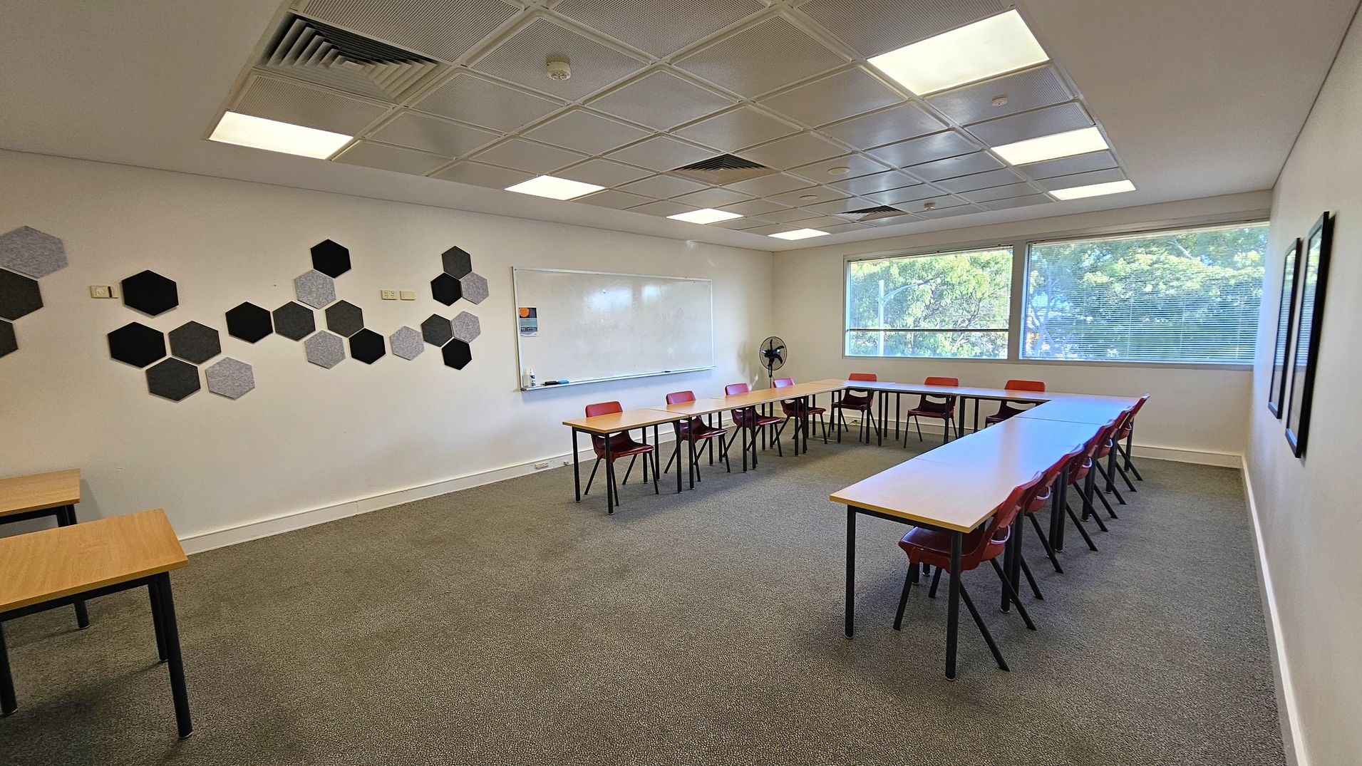 A classroom with a U-shaped arrangement of tables and red chairs. On the left wall, there are hexagonal acoustic panels in various shades of grey and black. The back wall features a large whiteboard. The right side has large windows with blinds, allowing natural light to enter. The ceiling is fitted with square lights and ventilation panels. The floor is carpeted in grey.