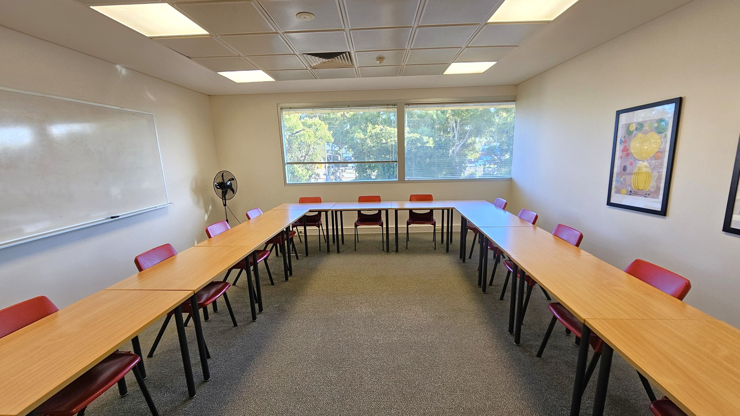 A classroom with U-shaped wooden tables and red chairs. On the left, a large whiteboard is mounted on the wall. The back wall features two large windows with a view of trees outside. A small fan is positioned near the windows. On the right wall, there are two framed abstract artworks. The ceiling has recessed lighting, and the floor is carpeted in a neutral tone.