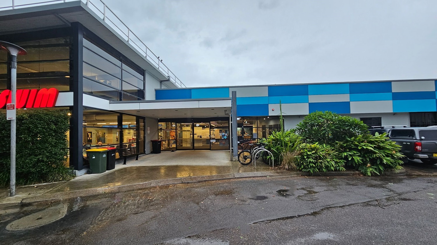 A car park with several vehicles is on the left, bordered by a grassy area and trees. On the right, a building with a blue and grey facade has large windows and a few parked cars in front. The ground is wet with puddles, and there are yellow bollards near the building. In the background, more trees and a grassy field are visible. A car park with several vehicles is on the left, bordered by a grassy area and trees. On the right, a building with a blue and grey facade has large windows and a few parked cars in front. The ground is wet with puddles, and there are yellow bollards near the building. In the background, more trees and a grassy field are visible.