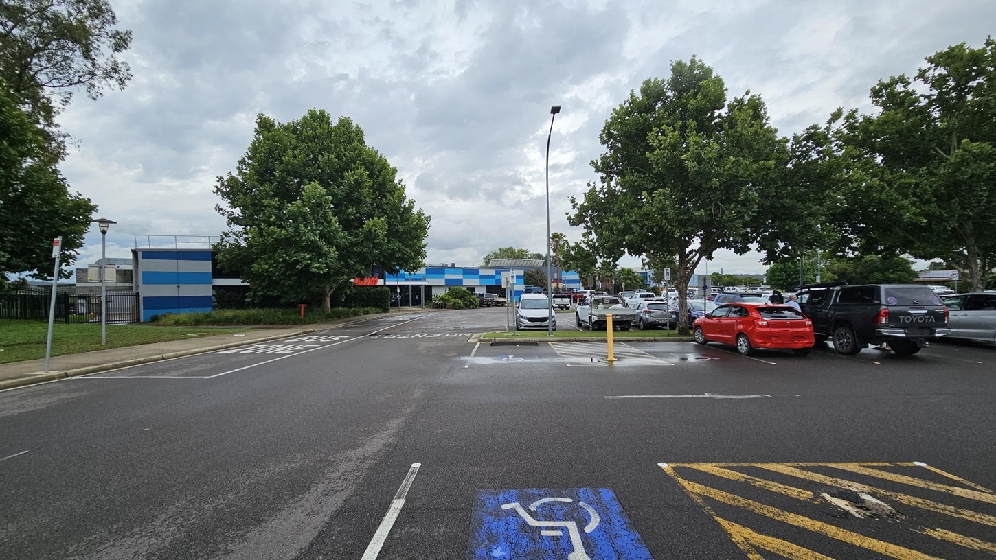 A car park with a blue and white building in the background. The foreground shows a disabled parking space marked in blue. On the right, several cars, including a red one and a black Toyota, are parked under trees. On the left, a footpath runs alongside a grassy area with a lamp post and more trees. The sky is overcast. A car park with a blue and white building in the background. The foreground shows a disabled parking space marked in blue. On the right, several cars, including a red one and a black Toyota, are parked under trees. On the left, a footpath runs alongside a grassy area with a lamp post and more trees. The sky is overcast.