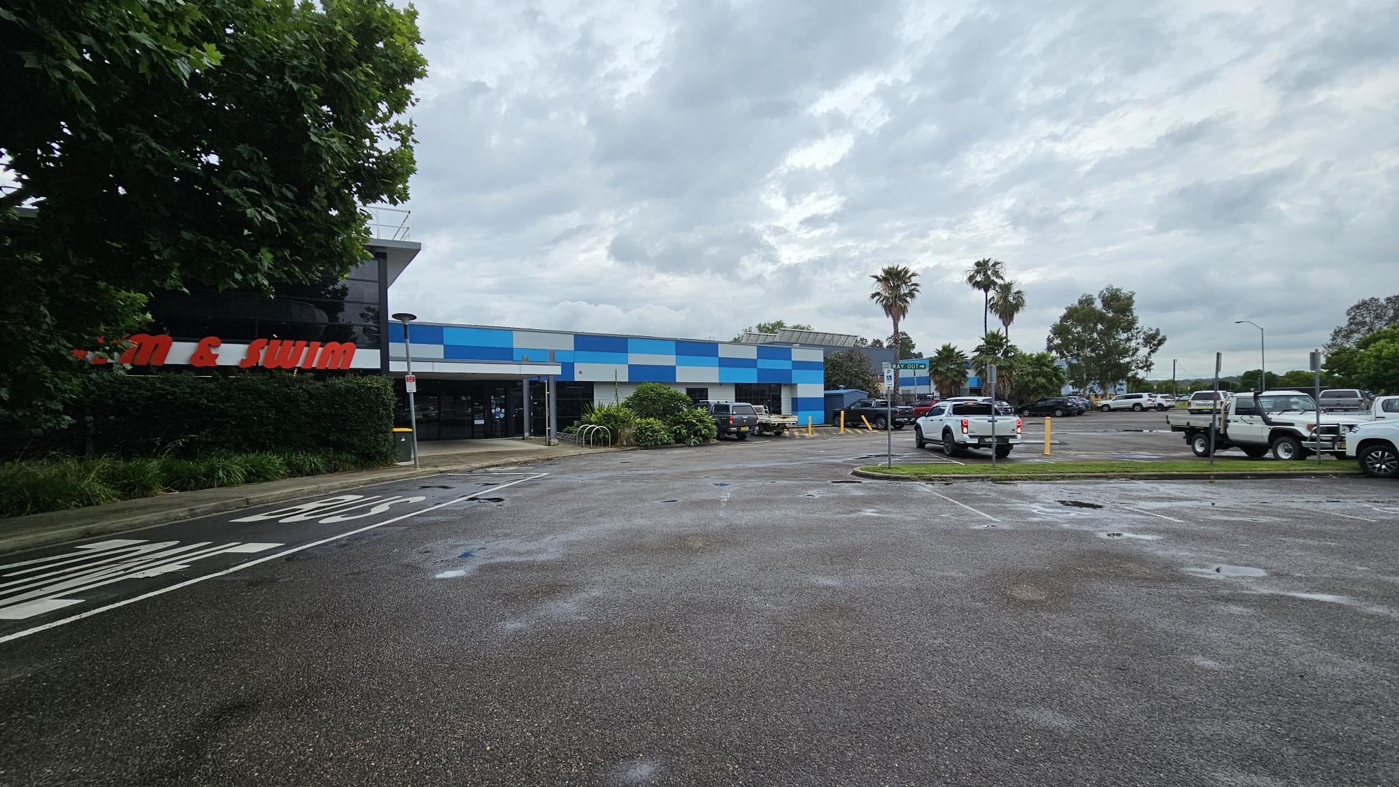 A car park with several vehicles, including white trucks, is in the foreground. On the left, a building with a partially visible red sign is surrounded by greenery. The building has a blue and white checkered pattern. In the background, there are palm trees and more parked cars. The sky is overcast. A car park with several vehicles, including white trucks, is in the foreground. On the left, a building with a partially visible red sign is surrounded by greenery. The building has a blue and white checkered pattern. In the background, there are palm trees and more parked cars. The sky is overcast.