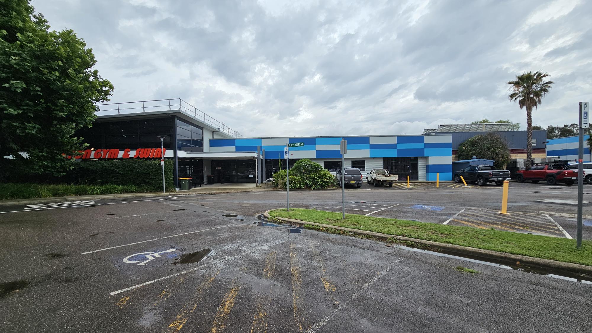 A parking area with marked spaces, including a disabled parking spot in the foreground. On the left, there's a building with large windows and a sign partially visible, surrounded by greenery. In the centre, a blue and white building with a sign above the entrance. Several vehicles are parked to the right, near a palm tree. The ground is wet, suggesting recent rain, and there are yellow bollards near the entrance. A parking area with marked spaces, including a disabled parking spot in the foreground. On the left, there's a building with large windows and a sign partially visible, surrounded by greenery. In the centre, a blue and white building with a sign above the entrance. Several vehicles are parked to the right, near a palm tree. The ground is wet, suggesting recent rain, and there are yellow bollards near the entrance.