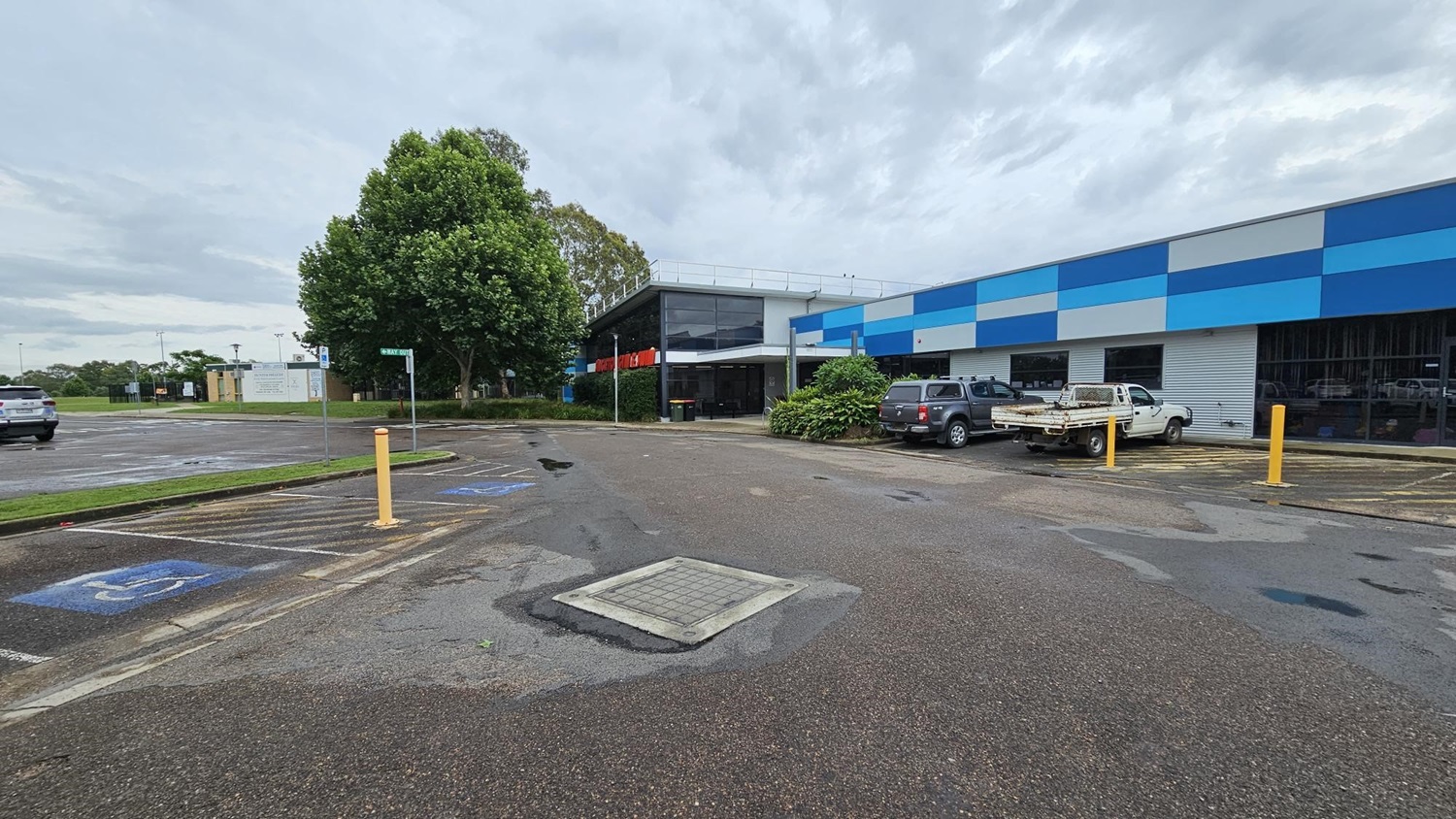 A car park with marked spaces, including disabled parking, is in the foreground. On the right, a building with a blue and white striped facade has several parked vehicles in front. In the centre, a small bush and a tree are near the building entrance. On the left, a large tree and a grassy area are visible, with a signpost nearby. The sky is overcast, and the ground appears wet. A car park with marked spaces, including disabled parking, is in the foreground. On the right, a building with a blue and white striped facade has several parked vehicles in front. In the centre, a small bush and a tree are near the building entrance. On the left, a large tree and a grassy area are visible, with a signpost nearby. The sky is overcast, and the ground appears wet.