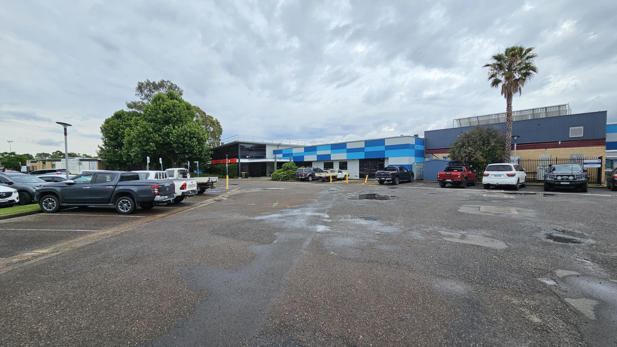 A car park with several vehicles, including a grey pickup and a white utility vehicle on the left. In the centre, a building with a blue and white checkered pattern is visible. On the right, a red car and a white SUV are parked near a palm tree. The ground is asphalt with some puddles, and there are trees and shrubs around the perimeter. A car park with several vehicles, including a grey pickup and a white utility vehicle on the left. In the centre, a building with a blue and white checkered pattern is visible. On the right, a red car and a white SUV are parked near a palm tree. The ground is asphalt with some puddles, and there are trees and shrubs around the perimeter.