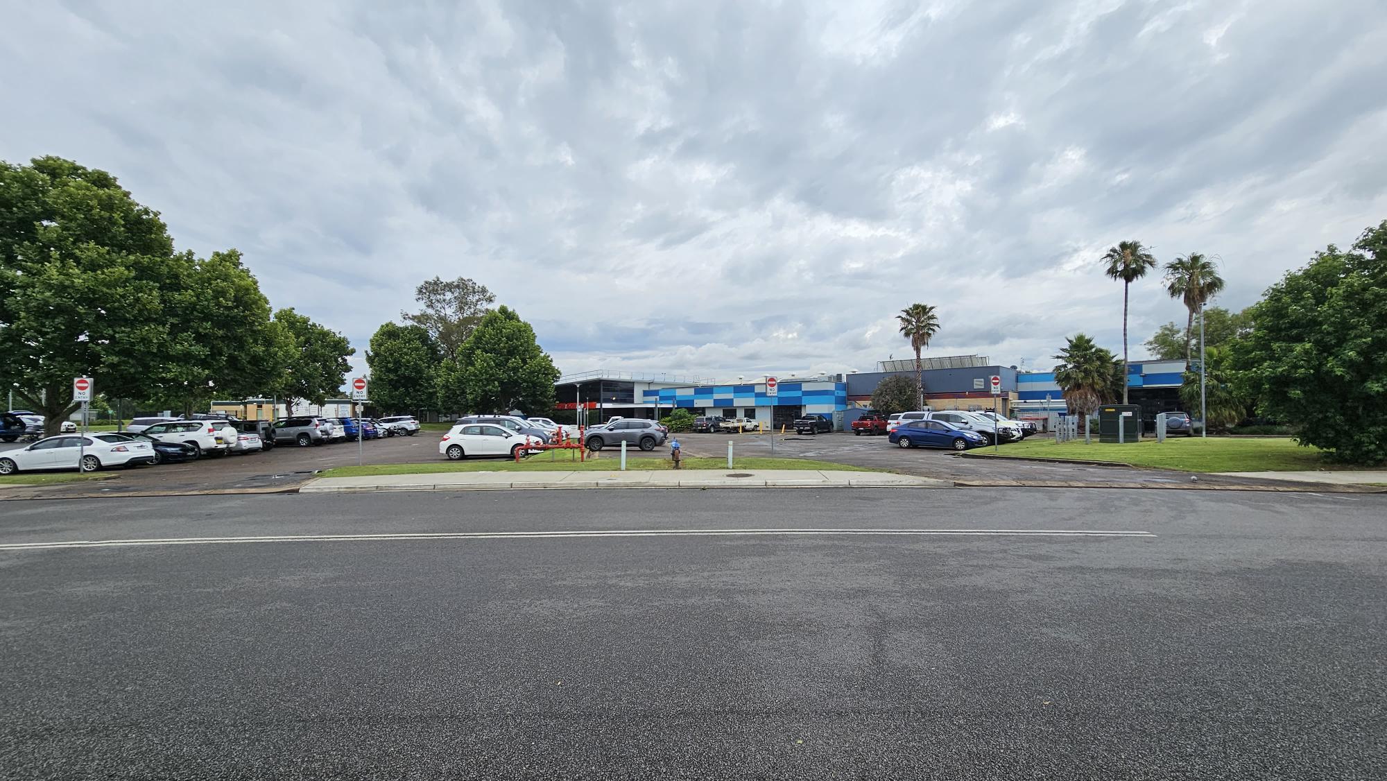 A car park with several vehicles is in the foreground, bordered by green grass and trees on both sides. In the background, there is a blue and grey industrial building. The sky is overcast, and there are palm trees near the building. The road in the foreground is empty, with a clear view of the parking area and the building beyond. A car park with several vehicles is in the foreground, bordered by green grass and trees on both sides. In the background, there is a blue and grey industrial building. The sky is overcast, and there are palm trees near the building. The road in the foreground is empty, with a clear view of the parking area and the building beyond.
