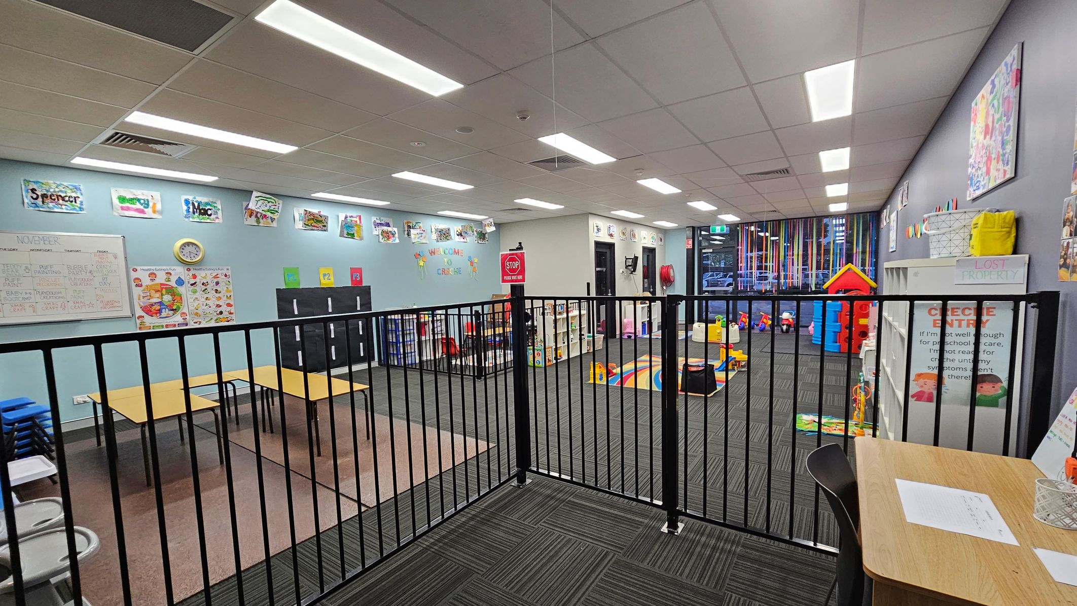 A brightly lit childcare room with a black safety gate in the foreground. On the left, there are tables and chairs, educational posters, and a clock on a light blue wall. The middle area has a colourful play mat and toys, with a "Welcome to Creche" sign above. On the right, shelves hold various items, and a small red playhouse is visible. The ceiling has recessed lighting, and the floor is carpeted in dark grey. A brightly lit childcare room with a black safety gate in the foreground. On the left, there are tables and chairs, educational posters, and a clock on a light blue wall. The middle area has a colourful play mat and toys, with a "Welcome to Creche" sign above. On the right, shelves hold various items, and a small red playhouse is visible. The ceiling has recessed lighting, and the floor is carpeted in dark grey.