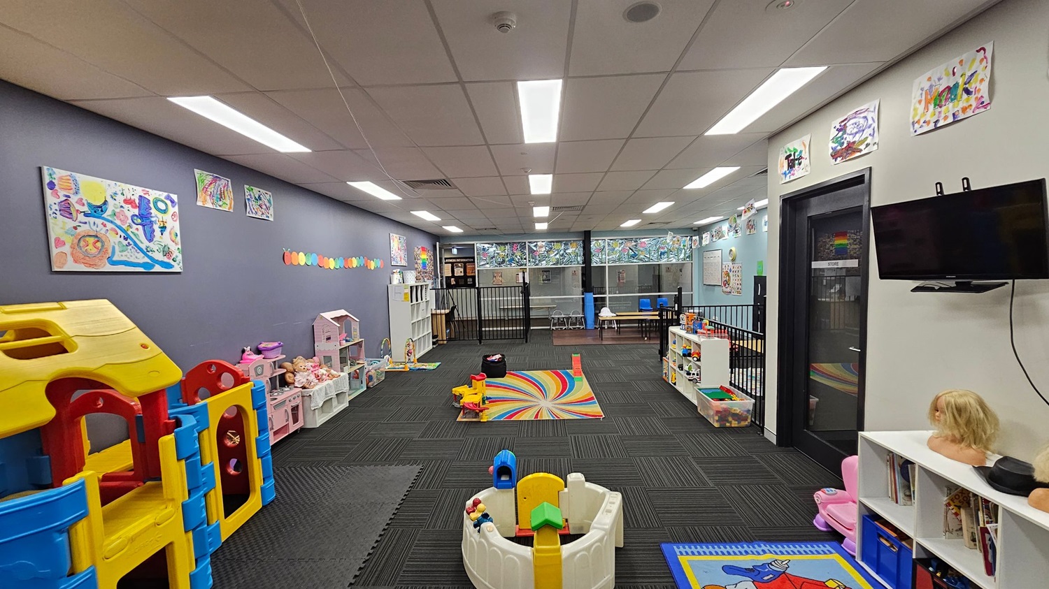 A colourful playroom with a variety of toys and activities. On the left, there's a large plastic play structure in yellow, red, and blue. Along the left wall are shelves with dolls and a dollhouse. The centre features a rainbow-patterned rug and various toys. On the right, a wall-mounted TV is above a door labelled "STORE". The walls are decorated with children's artwork. The floor is covered with dark carpet tiles, and the ceiling has bright fluorescent lights. A colourful playroom with a variety of toys and activities. On the left, there's a large plastic play structure in yellow, red, and blue. Along the left wall are shelves with dolls and a dollhouse. The centre features a rainbow-patterned rug and various toys. On the right, a wall-mounted TV is above a door labelled "STORE". The walls are decorated with children's artwork. The floor is covered with dark carpet tiles, and the ceiling has bright fluorescent lights.