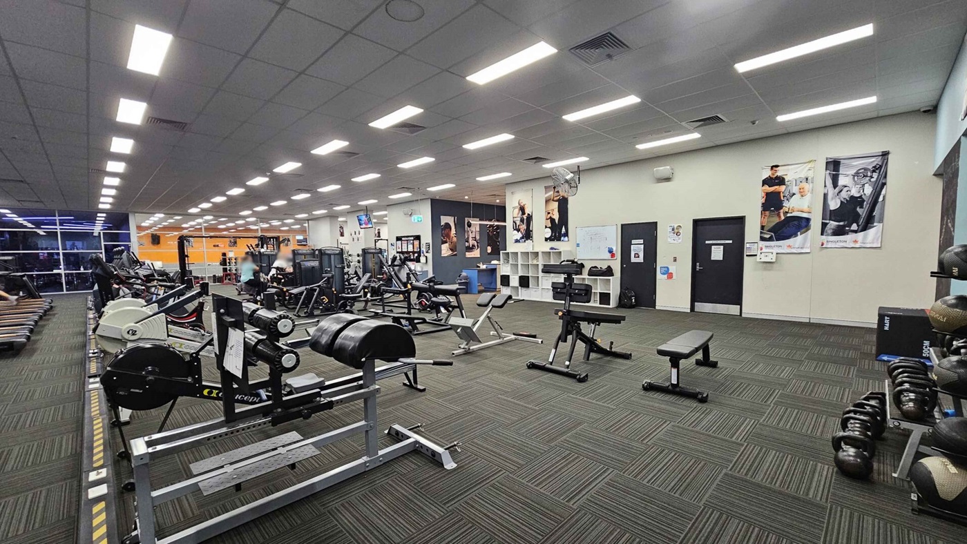 A spacious gym with various exercise equipment. On the left, there are rowing machines and treadmills near large windows. In the centre, weightlifting benches and machines are arranged neatly. The right side features kettlebells and medicine balls on racks. The walls have fitness posters, and there are two doors. The floor is covered with green and grey patterned carpet tiles, and the ceiling has bright fluorescent lights. A spacious gym with various exercise equipment. On the left, there are rowing machines and treadmills near large windows. In the centre, weightlifting benches and machines are arranged neatly. The right side features kettlebells and medicine balls on racks. The walls have fitness posters, and there are two doors. The floor is covered with green and grey patterned carpet tiles, and the ceiling has bright fluorescent lights.