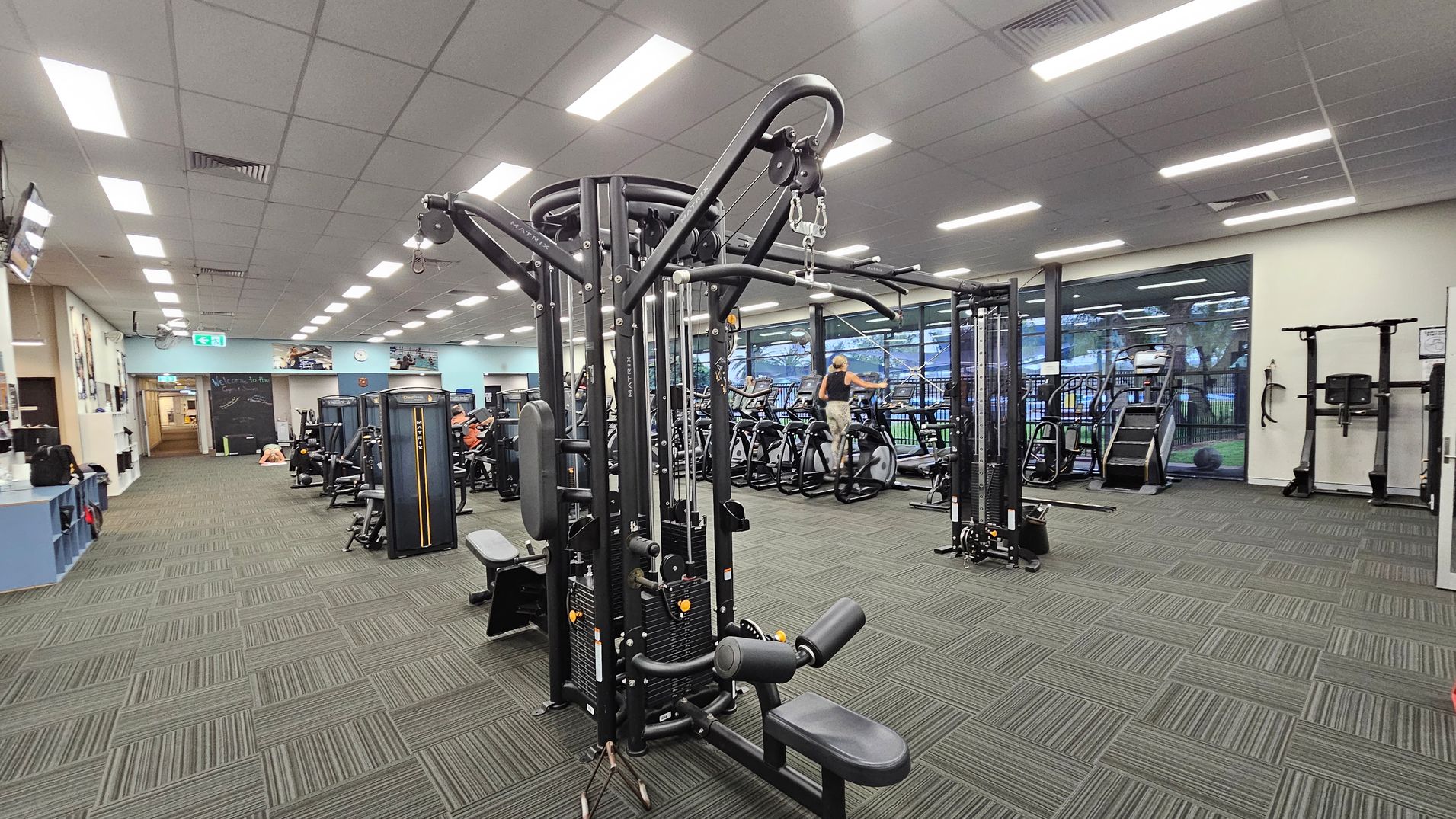 A spacious gym with various exercise machines. In the centre, there's a large multi-functional weight machine. To the right, there are treadmills and exercise bikes near large windows. The left side has a reception area with a counter and a chalkboard wall. The floor is covered with green and grey patterned carpet tiles, and the ceiling has bright fluorescent lights. A spacious gym with various exercise machines. In the centre, there's a large multi-functional weight machine. To the right, there are treadmills and exercise bikes near large windows. The left side has a reception area with a counter and a chalkboard wall. The floor is covered with green and grey patterned carpet tiles, and the ceiling has bright fluorescent lights.