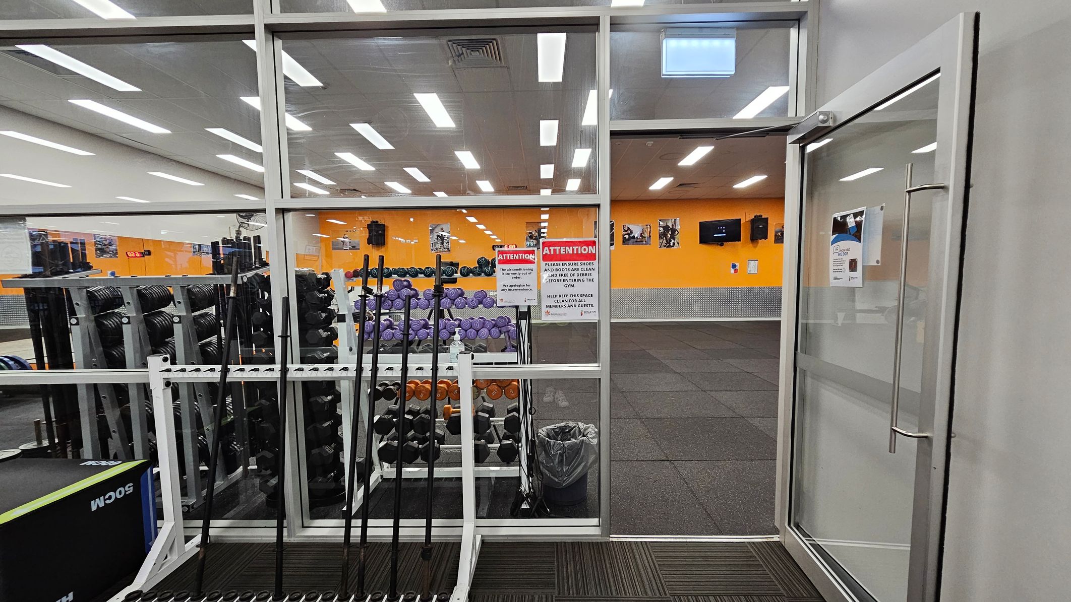 A gym interior with a glass wall and door on the right. Inside, there are racks of dumbbells and weightlifting bars. The walls are orange with several posters and a TV screen. The floor is covered with black rubber mats. Bright ceiling lights illuminate the space. Signs on the glass wall provide instructions for gym users. A trash bin is visible near the weights. A gym interior with a glass wall and door on the right. Inside, there are racks of dumbbells and weightlifting bars. The walls are orange with several posters and a TV screen. The floor is covered with black rubber mats. Bright ceiling lights illuminate the space. Signs on the glass wall provide instructions for gym users. A trash bin is visible near the weights.