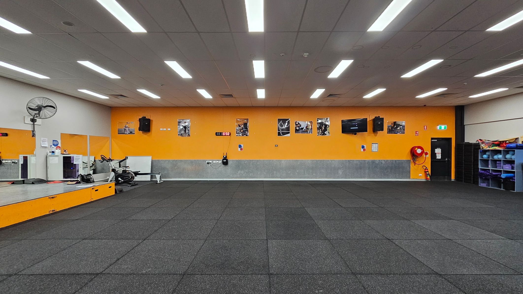 A gym room with a black rubber-tiled floor and a white ceiling with fluorescent lights. The back wall is orange with several fitness posters and a digital clock. On the left, there's a raised platform with a fan, exercise bike, and some equipment. On the right, a black door, fire extinguisher, and shelves with exercise mats and balls. The wall has a metallic panel running along its lower half. A gym room with a black rubber-tiled floor and a white ceiling with fluorescent lights. The back wall is orange with several fitness posters and a digital clock. On the left, there's a raised platform with a fan, exercise bike, and some equipment. On the right, a black door, fire extinguisher, and shelves with exercise mats and balls. The wall has a metallic panel running along its lower half.