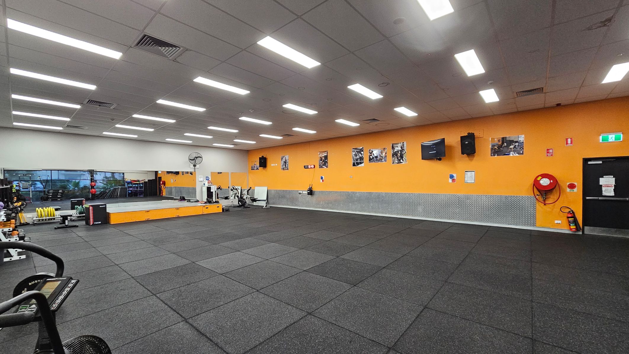 A spacious gym with a black rubber-tiled floor and a white ceiling with fluorescent lights. The left side features a large mirror and various gym equipment, including weights and exercise machines. The back wall is orange, adorned with several posters, a TV, and a speaker. A fire hose reel and an emergency exit sign are visible on the right. A raised platform is positioned near the mirror, and a fan is mounted on the wall. A spacious gym with a black rubber-tiled floor and a white ceiling with fluorescent lights. The left side features a large mirror and various gym equipment, including weights and exercise machines. The back wall is orange, adorned with several posters, a TV, and a speaker. A fire hose reel and an emergency exit sign are visible on the right. A raised platform is positioned near the mirror, and a fan is mounted on the wall.