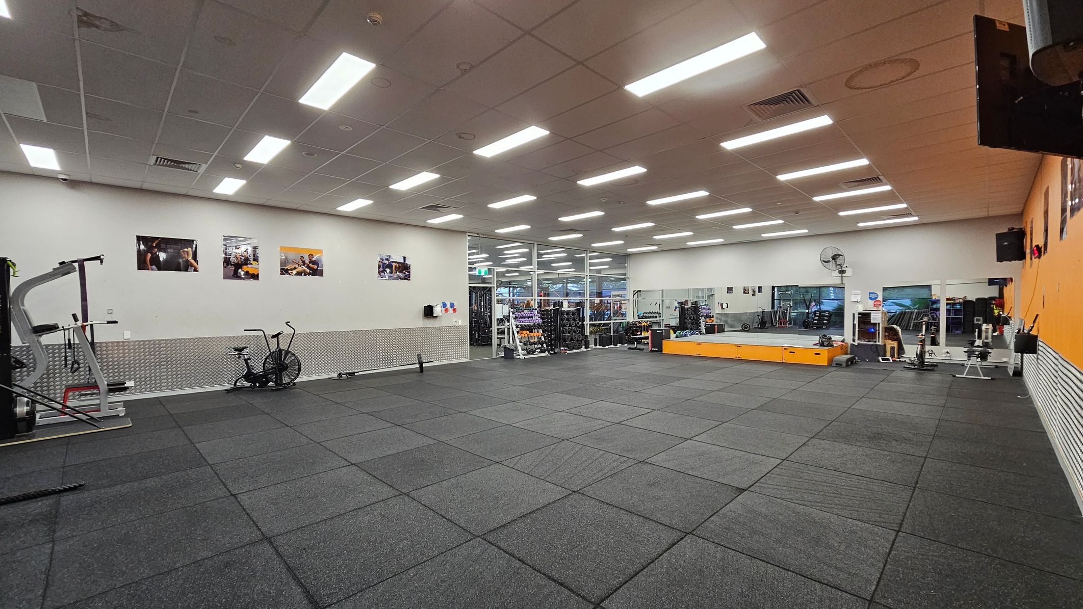 A spacious gym with a grey rubber floor and a white ceiling with fluorescent lights. On the left, there's exercise equipment including a stationary bike and a weight machine. The back wall features several framed photos and a pegboard with hanging items. The right side has a large mirror and a raised platform with more equipment. A fan is mounted on the wall, and a glass partition separates the gym from another area. A spacious gym with a grey rubber floor and a white ceiling with fluorescent lights. On the left, there's exercise equipment including a stationary bike and a weight machine. The back wall features several framed photos and a pegboard with hanging items. The right side has a large mirror and a raised platform with more equipment. A fan is mounted on the wall, and a glass partition separates the gym from another area.