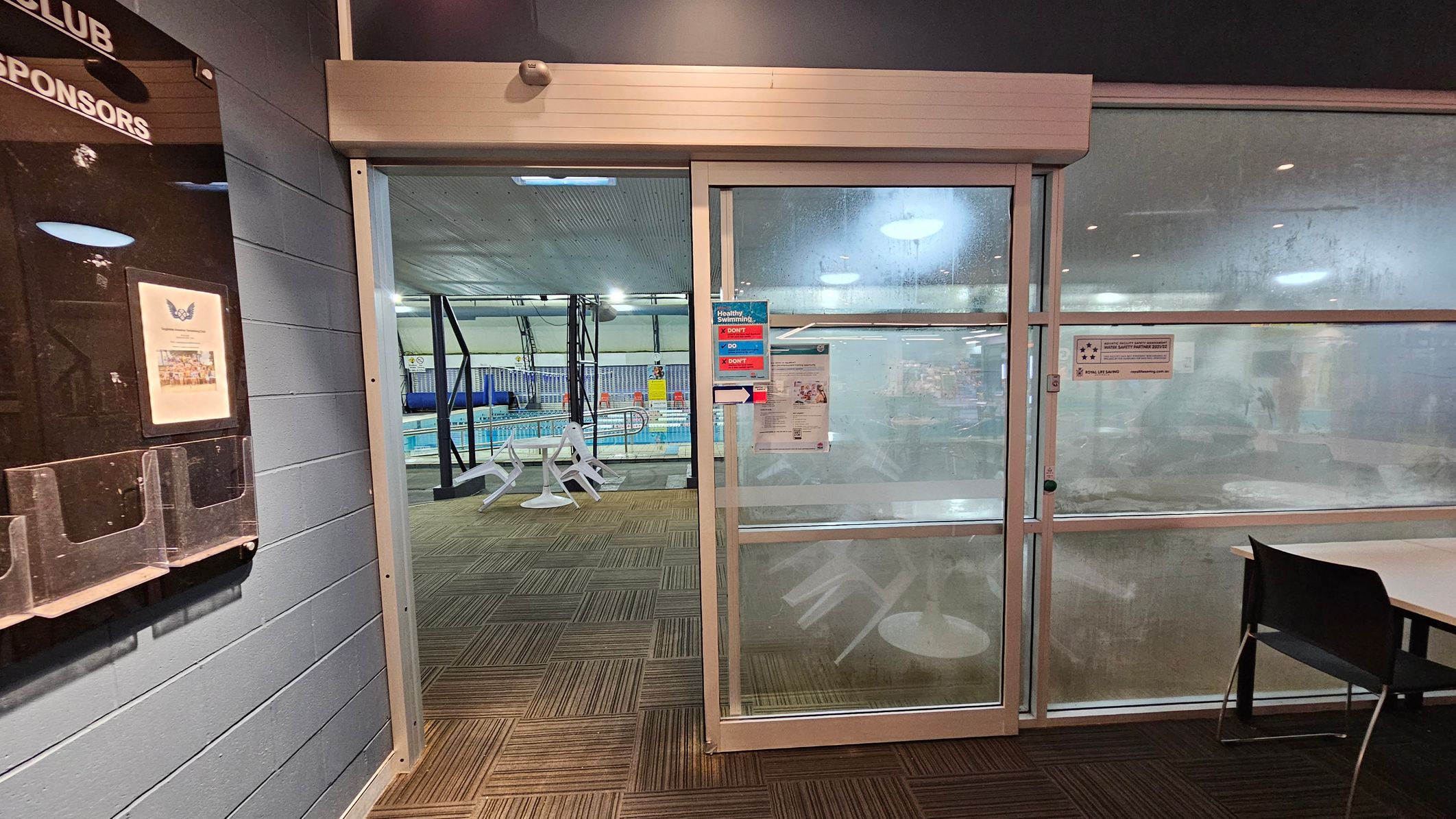 A glass door with a metal frame leads to an indoor swimming pool area. On the left, a notice board is mounted on a grey brick wall. The floor is covered with brown and green patterned carpet tiles. Through the glass, a pool with blue water and white poolside furniture is visible. On the right, a black chair is partially visible next to a white table. The ceiling is lined with lights, and there are signs and notices on the glass door. A glass door with a metal frame leads to an indoor swimming pool area. On the left, a notice board is mounted on a grey brick wall. The floor is covered with brown and green patterned carpet tiles. Through the glass, a pool with blue water and white poolside furniture is visible. On the right, a black chair is partially visible next to a white table. The ceiling is lined with lights, and there are signs and notices on the glass door.
