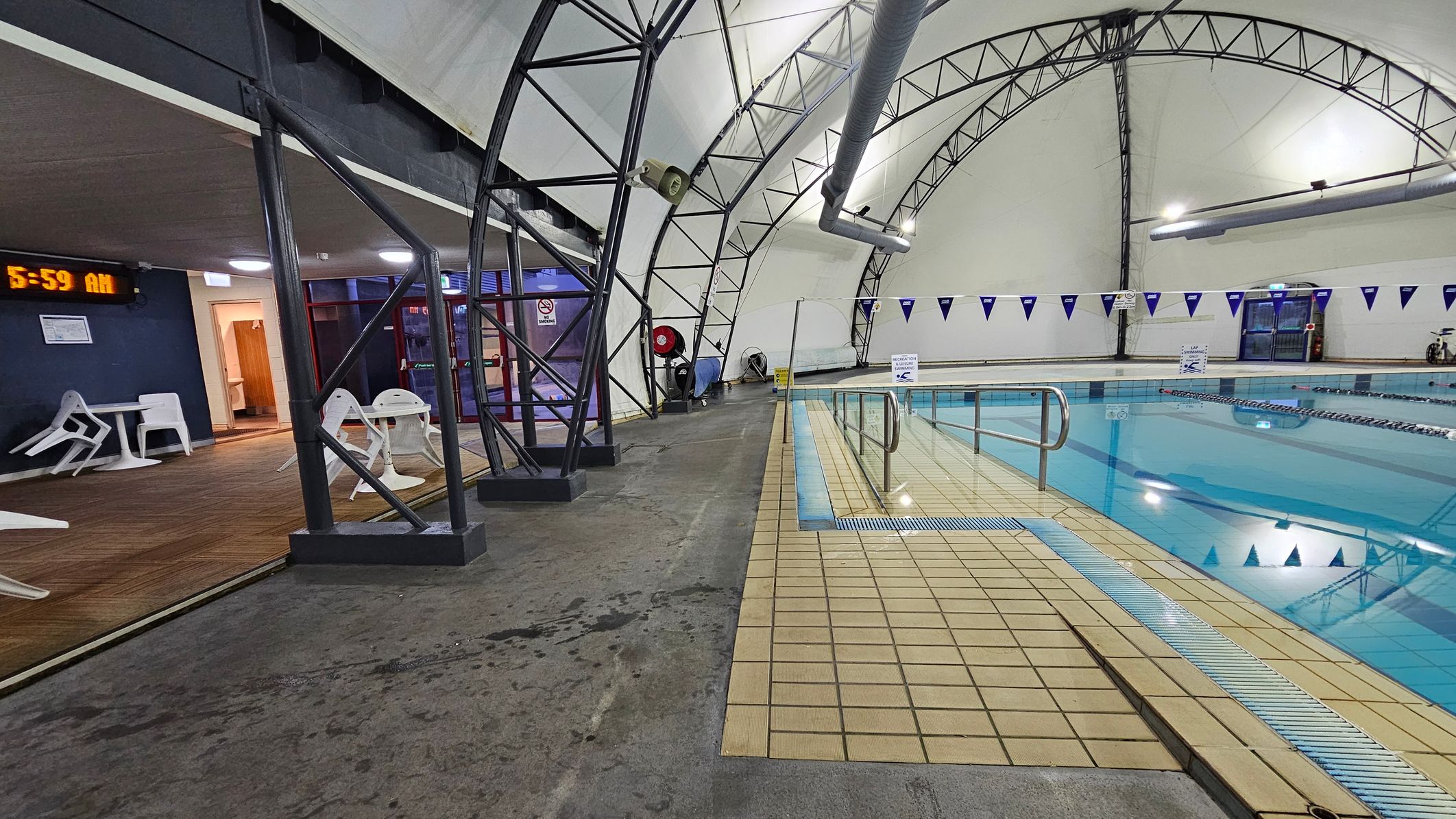 An indoor swimming pool with clear blue water is on the right, bordered by beige tiles. A metal handrail leads into the pool. On the left, there is a seating area with white plastic chairs and tables on a wooden floor. A digital clock on the wall reads "5:59 AM". The ceiling is a curved structure with metal beams and large ventilation pipes. Blue triangular flags hang above the pool. The floor is a mix of concrete and tile. An indoor swimming pool with clear blue water is on the right, bordered by beige tiles. A metal handrail leads into the pool. On the left, there is a seating area with white plastic chairs and tables on a wooden floor. A digital clock on the wall reads "5:59 AM". The ceiling is a curved structure with metal beams and large ventilation pipes. Blue triangular flags hang above the pool. The floor is a mix of concrete and tile.