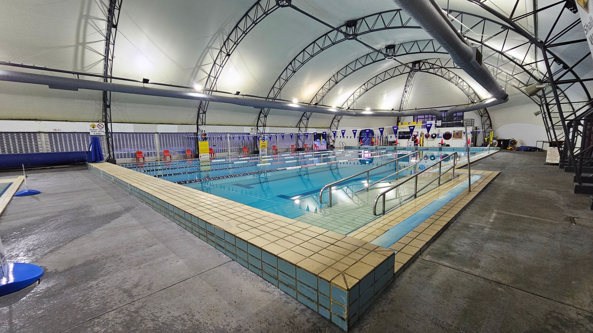 An indoor swimming pool with clear blue water, surrounded by a tiled deck. On the left, there is a wet concrete floor. The pool has metal handrails and lane dividers. The ceiling is a curved, white structure supported by metal beams. Along the far wall, there are red chairs and various signs. The right side has more concrete flooring and some equipment. The lighting is bright, illuminating the entire space. An indoor swimming pool with clear blue water, surrounded by a tiled deck. On the left, there is a wet concrete floor. The pool has metal handrails and lane dividers. The ceiling is a curved, white structure supported by metal beams. Along the far wall, there are red chairs and various signs. The right side has more concrete flooring and some equipment. The lighting is bright, illuminating the entire space.