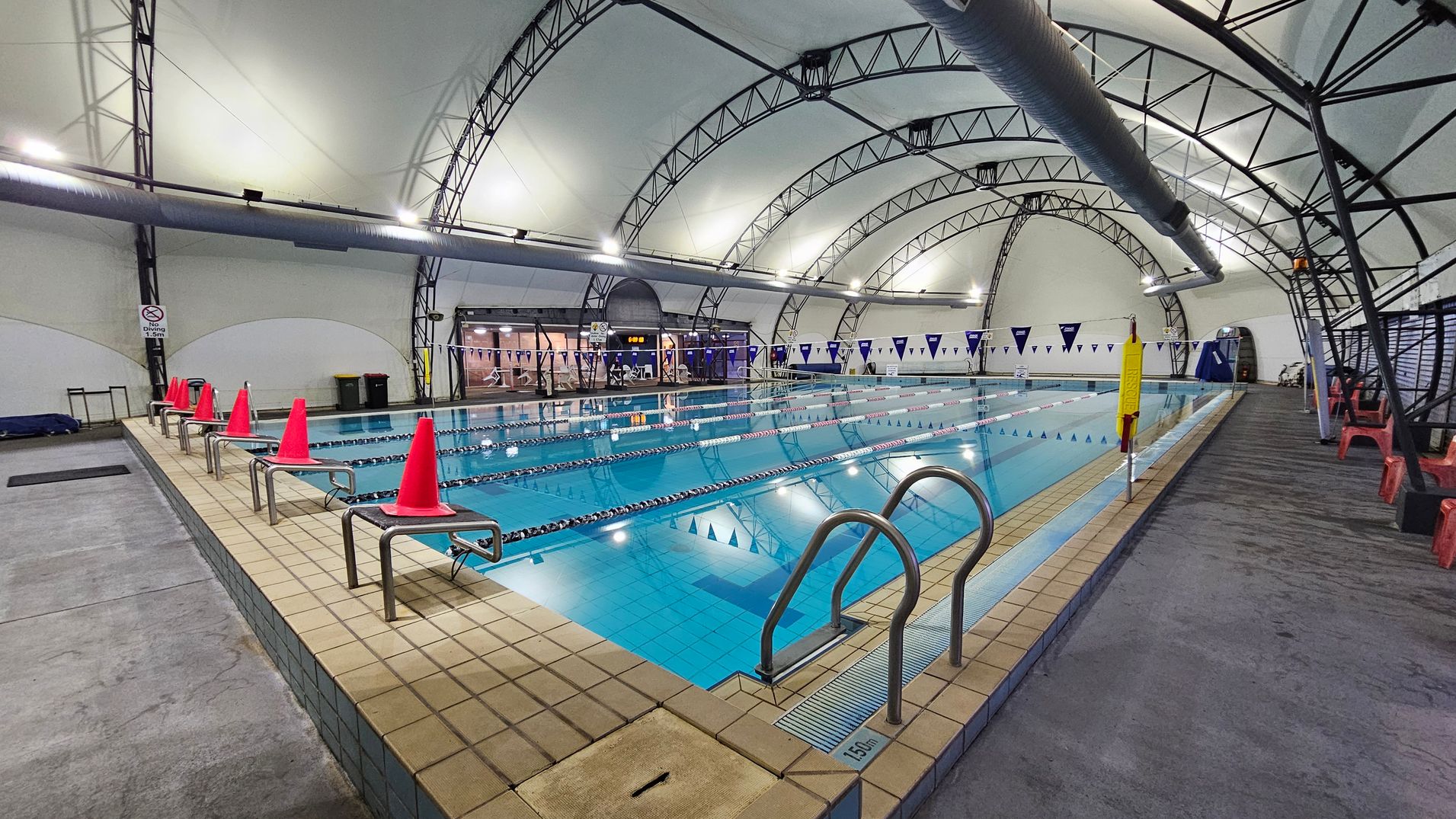 An indoor swimming pool with clear blue water, divided into lanes by floating ropes. On the left, a row of red traffic cones lines the pool's edge. The right side has a series of red plastic chairs on a grey concrete floor. The ceiling is a curved structure with metal beams and large ventilation ducts. In the background, a glass entrance with a digital clock above it is visible, along with blue and white triangular flags hanging across the pool. An indoor swimming pool with clear blue water, divided into lanes by floating ropes. On the left, a row of red traffic cones lines the pool's edge. The right side has a series of red plastic chairs on a grey concrete floor. The ceiling is a curved structure with metal beams and large ventilation ducts. In the background, a glass entrance with a digital clock above it is visible, along with blue and white triangular flags hanging across the pool.