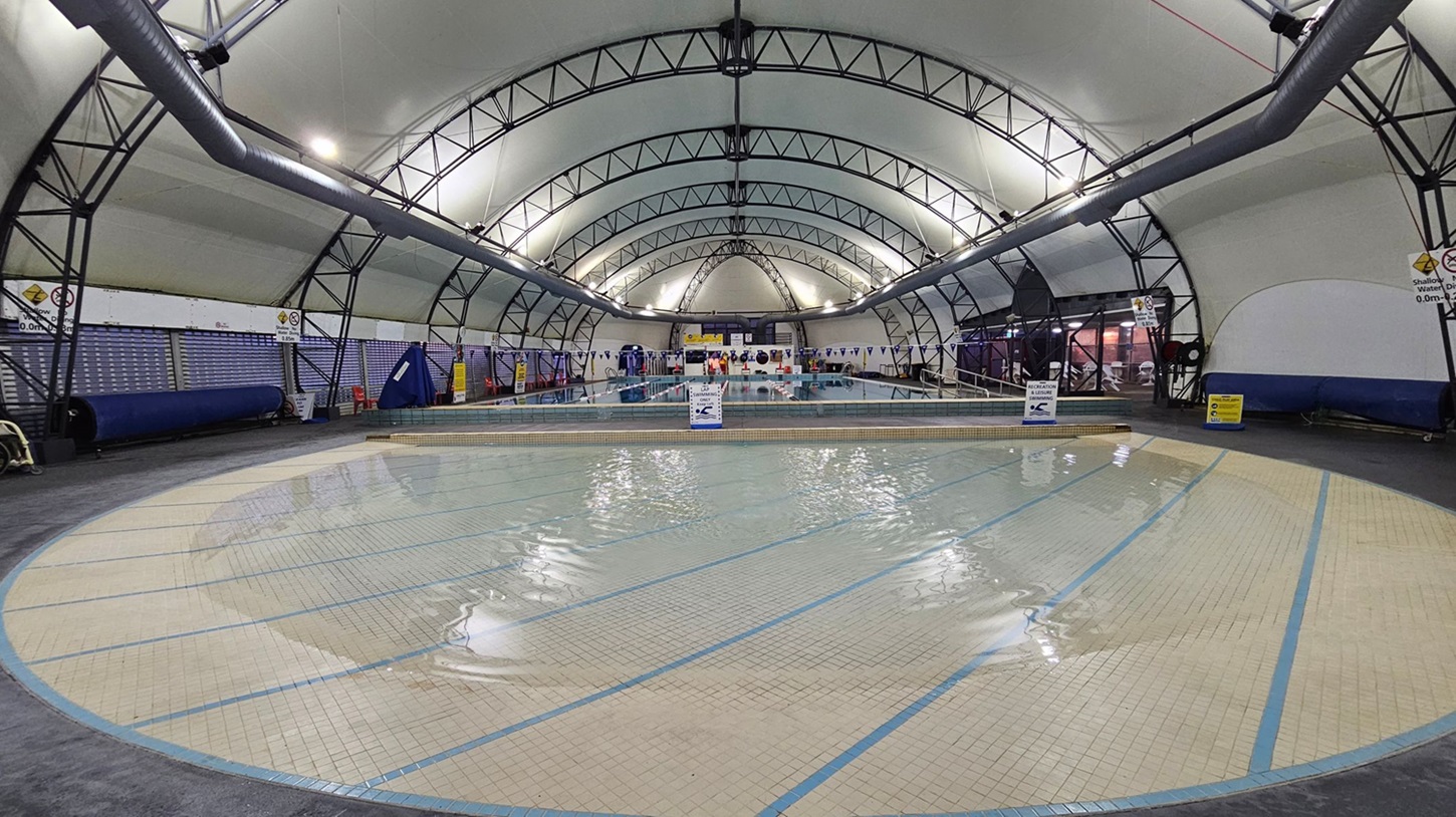 An indoor swimming pool with a shallow tiled area in the foreground, featuring light green and beige tiles. The pool is in the centre, surrounded by a wide deck. The ceiling is a high, arched structure with metal beams and large ventilation ducts running along both sides. Signs and equipment are visible along the walls. In the background, there are people near the pool, and the area is well-lit with overhead lights. An indoor swimming pool with a shallow tiled area in the foreground, featuring light green and beige tiles. The pool is in the centre, surrounded by a wide deck. The ceiling is a high, arched structure with metal beams and large ventilation ducts running along both sides. Signs and equipment are visible along the walls. In the background, there are people near the pool, and the area is well-lit with overhead lights.