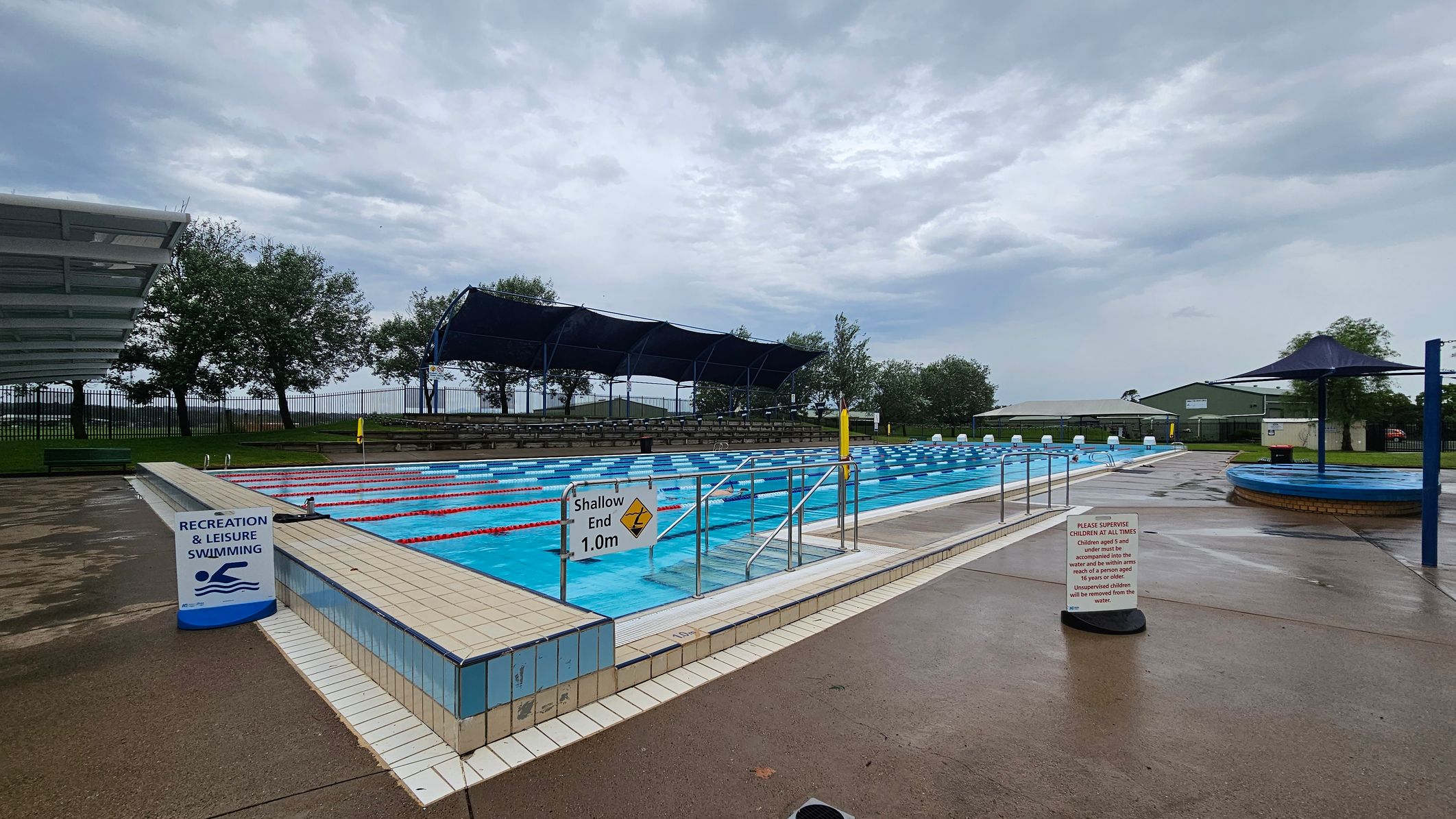 An outdoor swimming pool area with a wet concrete surface. On the left, there's a grassy area with a palm tree and a white pergola. The pool is in the centre, surrounded by a low fence, with blue and white lane dividers. A sign indicates a depth of 1.0m. On the right, there are blue shade structures and a small building in the background. Trees and a fence line the perimeter. An outdoor swimming pool area with a wet concrete surface. On the left, there's a grassy area with a palm tree and a white pergola. The pool is in the centre, surrounded by a low fence, with blue and white lane dividers. A sign indicates a depth of 1.0m. On the right, there are blue shade structures and a small building in the background. Trees and a fence line the perimeter.
