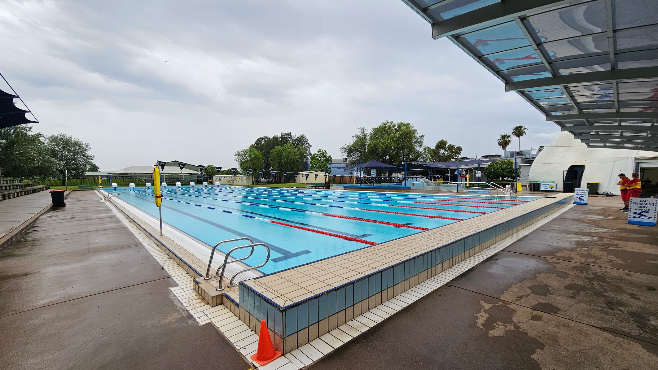 An outdoor swimming pool with multiple lanes, marked by red and blue lane dividers. On the left, there is a wet concrete walkway with a black bin and a lifebuoy. On the right, a covered area with a transparent roof and two people in red uniforms standing near signs that read "Lap Swimming." In the background, there are trees, a fence, and some buildings. The pool's edge is tiled, and a small orange cone is placed at the corner. An outdoor swimming pool with multiple lanes, marked by red and blue lane dividers. On the left, there is a wet concrete walkway with a black bin and a lifebuoy. On the right, a covered area with a transparent roof and two people in red uniforms standing near signs that read "Lap Swimming." In the background, there are trees, a fence, and some buildings. The pool's edge is tiled, and a small orange cone is placed at the corner.