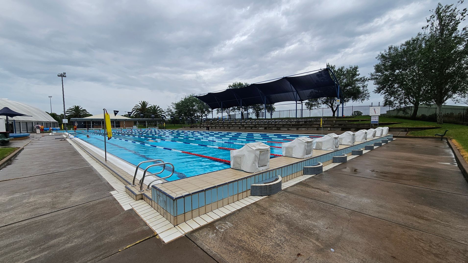 Outdoor swimming pool with multiple lanes, surrounded by a wet concrete deck. On the right, there are covered starting blocks and a grassy area with trees and a fence. On the left, a pathway leads to a building with a dome-shaped roof and palm trees nearby. A row of shaded seating is visible in the background. The pool has blue and white lane dividers, and a metal ladder is at the pool's edge. The sky is overcast. Outdoor swimming pool with multiple lanes, surrounded by a wet concrete deck. On the right, there are covered starting blocks and a grassy area with trees and a fence. On the left, a pathway leads to a building with a dome-shaped roof and palm trees nearby. A row of shaded seating is visible in the background. The pool has blue and white lane dividers, and a metal ladder is at the pool's edge. The sky is overcast.