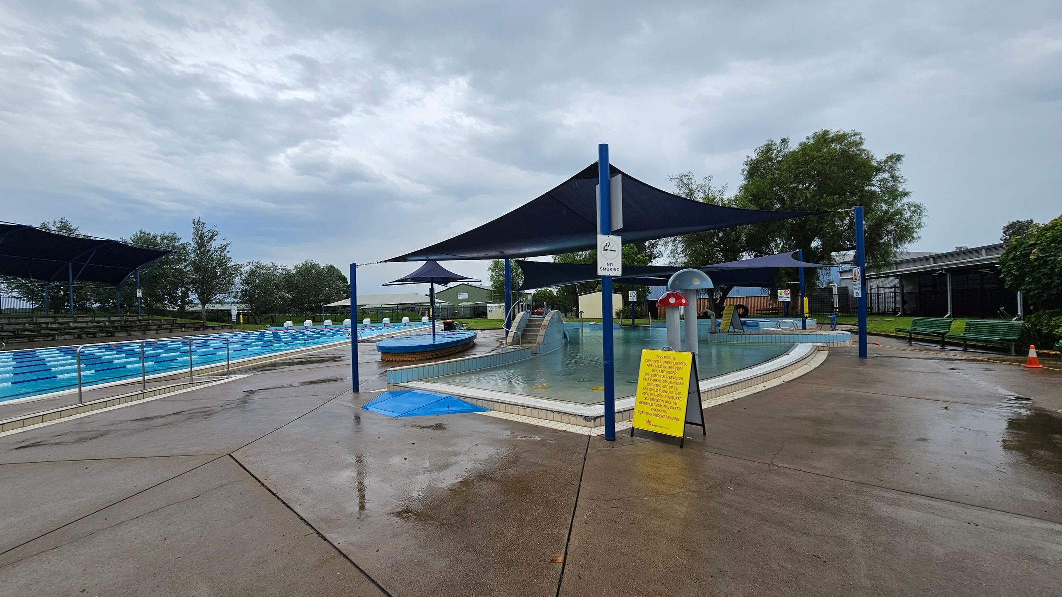 A swimming pool area with a wet concrete floor. On the left, there is a large pool with blue and white lane dividers, and a shaded seating area with trees in the background. In the centre, a smaller pool features a slide and a mushroom-shaped water feature, covered by blue shade sails. A yellow sign with pool rules is visible. On the right, there are green benches and more trees, with a building partially visible in the background. A swimming pool area with a wet concrete floor. On the left, there is a large pool with blue and white lane dividers, and a shaded seating area with trees in the background. In the centre, a smaller pool features a slide and a mushroom-shaped water feature, covered by blue shade sails. A yellow sign with pool rules is visible. On the right, there are green benches and more trees, with a building partially visible in the background.
