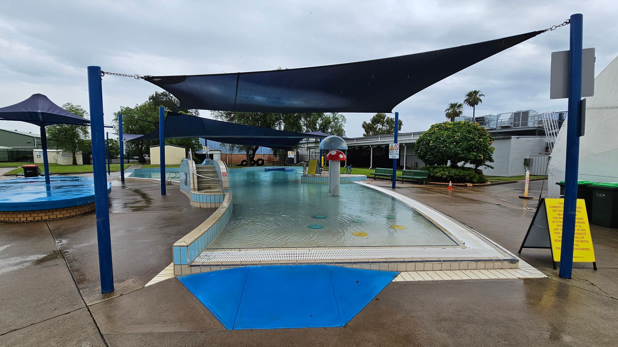 A children's splash pool area with a shallow water feature in the centre, surrounded by a tiled edge. A mushroom-shaped water fountain is visible in the middle. Blue shade sails are suspended above, supported by blue poles. To the right, a yellow sign is placed on the wet concrete ground. In the background, there are trees, a building with metal siding, and a grassy area with a bush. The sky is overcast. A children's splash pool area with a shallow water feature in the centre, surrounded by a tiled edge. A mushroom-shaped water fountain is visible in the middle. Blue shade sails are suspended above, supported by blue poles. To the right, a yellow sign is placed on the wet concrete ground. In the background, there are trees, a building with metal siding, and a grassy area with a bush. The sky is overcast.