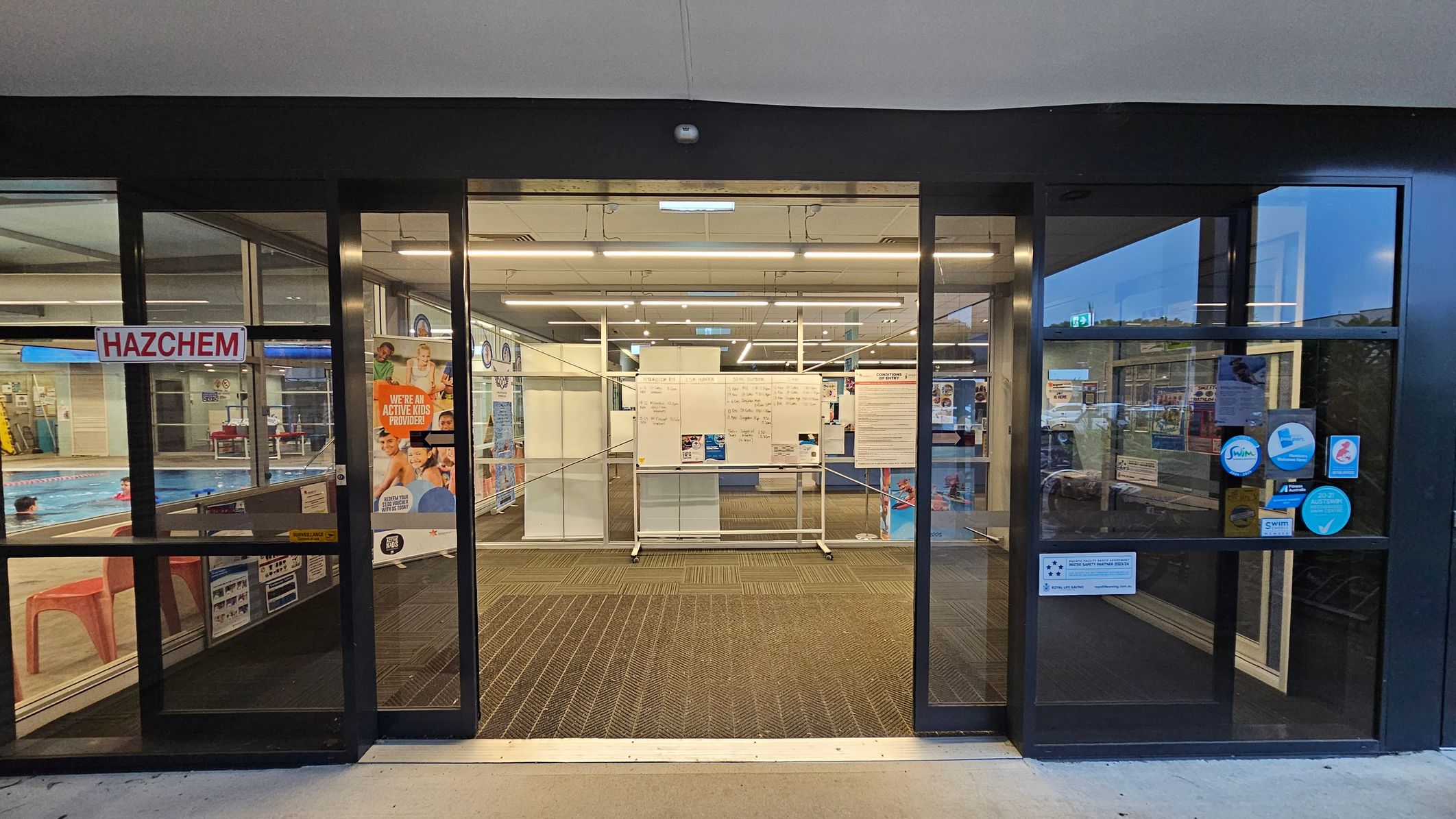 Glass entrance doors to a swimming pool facility. On the left, a "HAZCHEM" sign is visible, with a glimpse of the pool and swimmers inside. On the right, several stickers and notices are displayed on the glass. In the centre, a whiteboard with information is positioned behind the doors. The floor is carpeted with a patterned mat, and the ceiling has fluorescent lighting. Posters and promotional materials are visible inside. Glass entrance doors to a swimming pool facility. On the left, a "HAZCHEM" sign is visible, with a glimpse of the pool and swimmers inside. On the right, several stickers and notices are displayed on the glass. In the centre, a whiteboard with information is positioned behind the doors. The floor is carpeted with a patterned mat, and the ceiling has fluorescent lighting. Posters and promotional materials are visible inside.