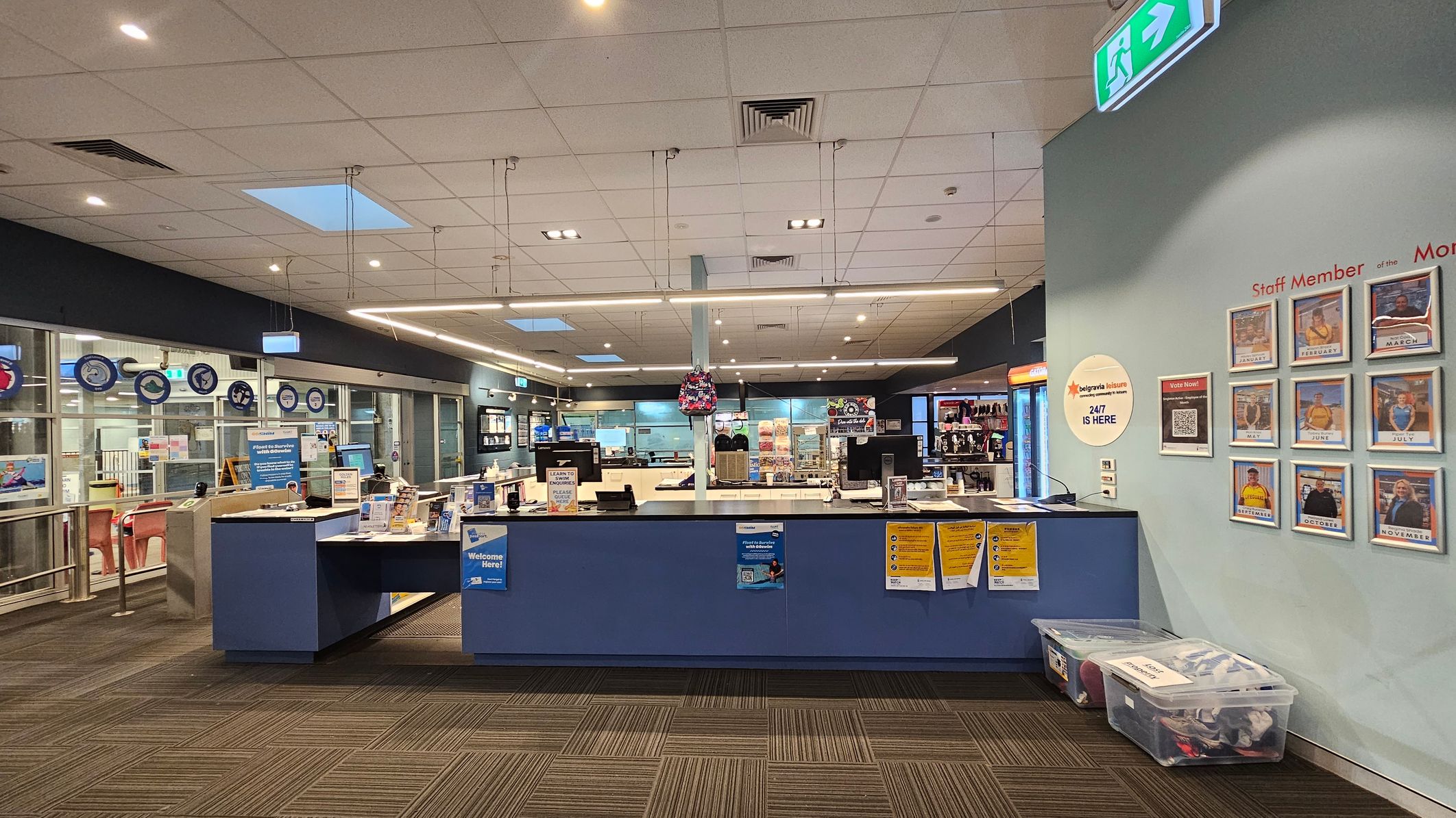 A reception area with a blue counter in the centre, displaying various informational materials and a small backpack hanging above. Behind the counter are computer monitors and a well-lit interior with shelves and displays. On the right wall, there are framed photos labelled with months, titled "Staff Member of the Month." Below, clear plastic bins are placed on the floor. The ceiling is fitted with lights and ventilation panels, and the floor has a patterned carpet. A reception area with a blue counter in the centre, displaying various informational materials and a small backpack hanging above. Behind the counter are computer monitors and a well-lit interior with shelves and displays. On the right wall, there are framed photos labelled with months, titled "Staff Member of the Month." Below, clear plastic bins are placed on the floor. The ceiling is fitted with lights and ventilation panels, and the floor has a patterned carpet.