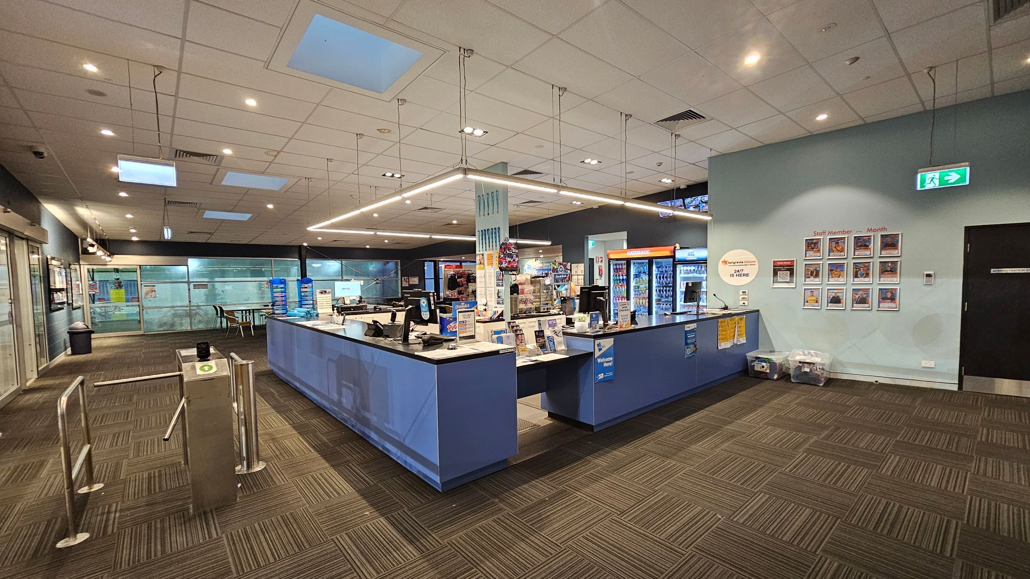 A spacious interior with a reception desk in the centre, featuring computers and promotional materials. To the right, a wall displays "Staff Member of the Month" photos and an emergency exit sign. On the left, a turnstile and glass doors lead to another area. The floor is carpeted with a geometric pattern, and the ceiling has recessed lighting and skylights. Vending machines and seating are visible in the background. A spacious interior with a reception desk in the centre, featuring computers and promotional materials. To the right, a wall displays "Staff Member of the Month" photos and an emergency exit sign. On the left, a turnstile and glass doors lead to another area. The floor is carpeted with a geometric pattern, and the ceiling has recessed lighting and skylights. Vending machines and seating are visible in the background.