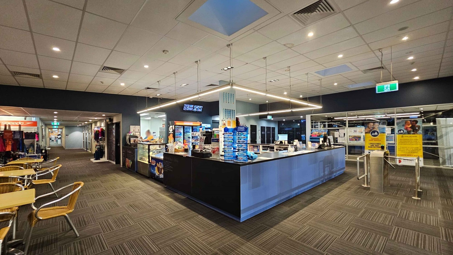 A spacious indoor area with a modern reception counter in the centre, displaying various products and a cash register. To the left, there are tables with wicker chairs and a display of clothing. The floor is carpeted with a geometric pattern. The ceiling is fitted with recessed lighting and a skylight. On the right, there are large glass windows with posters and a turnstile entrance. The background features a corridor leading to other areas. A spacious indoor area with a modern reception counter in the centre, displaying various products and a cash register. To the left, there are tables with wicker chairs and a display of clothing. The floor is carpeted with a geometric pattern. The ceiling is fitted with recessed lighting and a skylight. On the right, there are large glass windows with posters and a turnstile entrance. The background features a corridor leading to other areas.