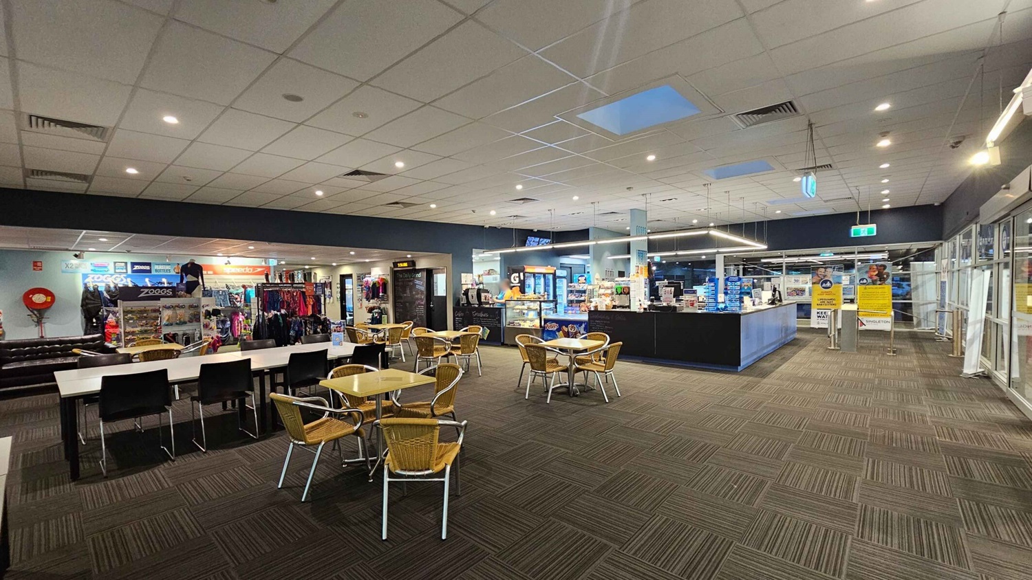 A spacious indoor area with a tiled ceiling and carpeted floor. In the foreground, there are tables with wicker chairs. On the left, a shop displays swimwear and accessories with brands like Zoggs and Speedo. In the centre, a café counter offers snacks and drinks. On the right, large glass doors and windows lead to another area, with signs and notices visible. The lighting is bright and evenly distributed. A spacious indoor area with a tiled ceiling and carpeted floor. In the foreground, there are tables with wicker chairs. On the left, a shop displays swimwear and accessories with brands like Zoggs and Speedo. In the centre, a café counter offers snacks and drinks. On the right, large glass doors and windows lead to another area, with signs and notices visible. The lighting is bright and evenly distributed.