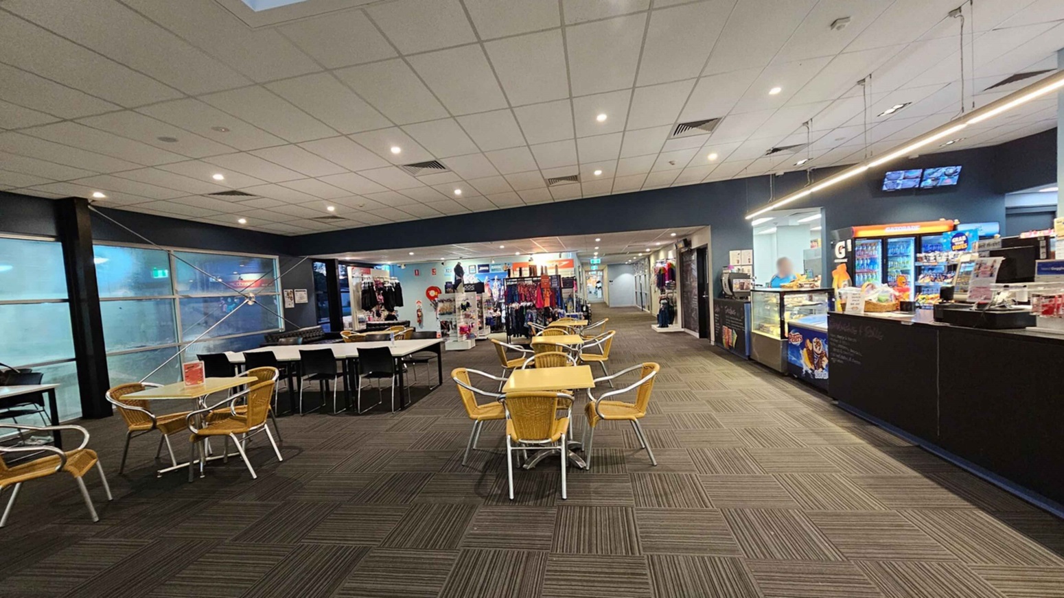 A spacious indoor area with a café on the right, featuring a counter displaying snacks and drinks. A person is behind the counter. In the centre, there are tables and chairs with wicker seats. On the left, a row of tables with black chairs is positioned near large frosted windows. In the background, a shop displays clothing and accessories. The ceiling is fitted with recessed lighting, and the floor is covered with patterned carpet tiles. A spacious indoor area with a café on the right, featuring a counter displaying snacks and drinks. A person is behind the counter. In the centre, there are tables and chairs with wicker seats. On the left, a row of tables with black chairs is positioned near large frosted windows. In the background, a shop displays clothing and accessories. The ceiling is fitted with recessed lighting, and the floor is covered with patterned carpet tiles.