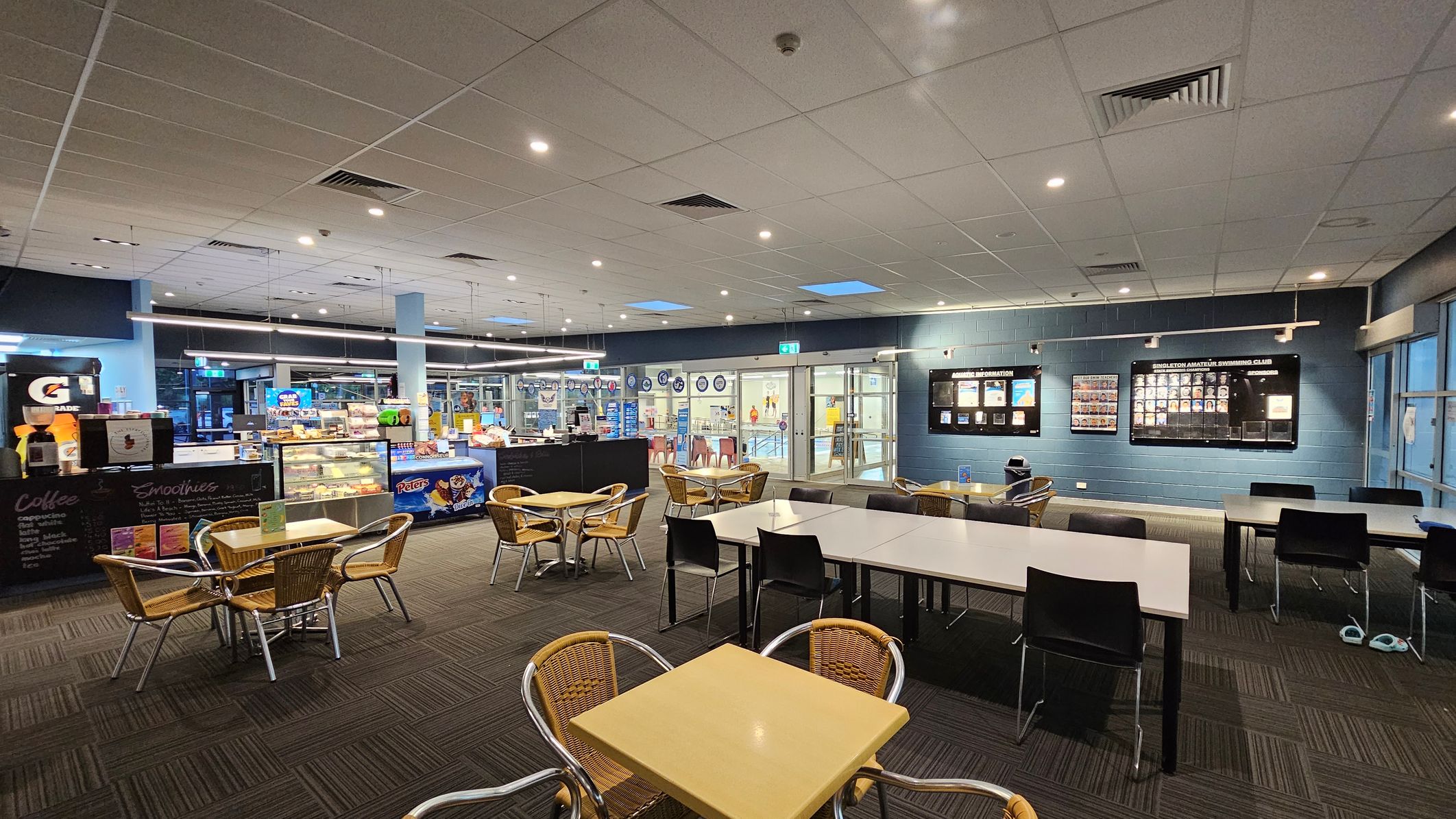 A spacious indoor café area with a tiled ceiling and carpeted floor. On the left, there is a counter displaying snacks and drinks, with a Gatorade dispenser and a menu board for coffee and smoothies. In the centre, several tables with wicker chairs are arranged. On the right, a wall features display boards with information and photos. The background shows glass doors leading to another area, possibly a pool or gym. A spacious indoor café area with a tiled ceiling and carpeted floor. On the left, there is a counter displaying snacks and drinks, with a Gatorade dispenser and a menu board for coffee and smoothies. In the centre, several tables with wicker chairs are arranged. On the right, a wall features display boards with information and photos. The background shows glass doors leading to another area, possibly a pool or gym.