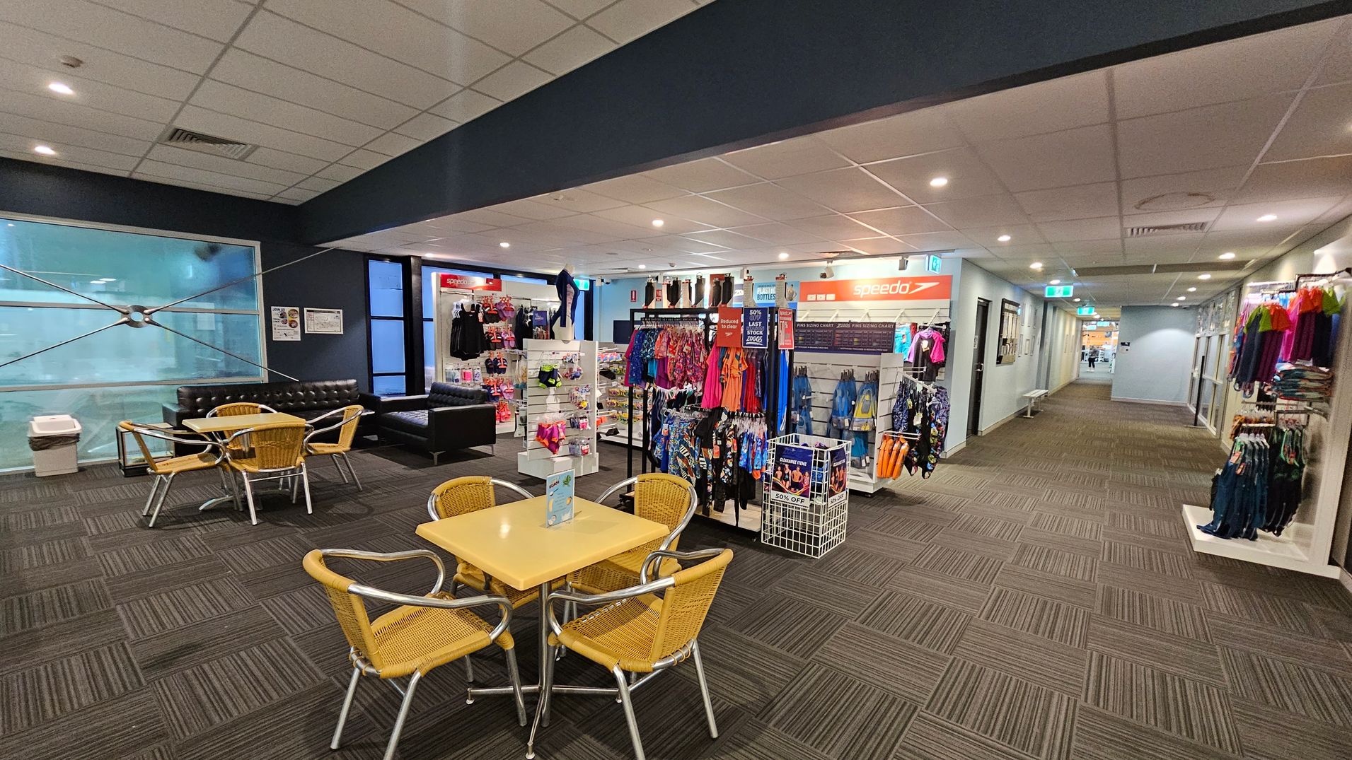 A retail area with a seating section on the left, featuring a yellow table and wicker chairs. Behind the seating, there are black leather sofas and a large window. In the centre, there are displays of swimwear and accessories, with racks of colourful clothing and a Speedo sign above. On the right, a corridor leads further into the building, with more clothing racks along the wall. The floor is carpeted with a striped pattern, and the ceiling has recessed lighting. A retail area with a seating section on the left, featuring a yellow table and wicker chairs. Behind the seating, there are black leather sofas and a large window. In the centre, there are displays of swimwear and accessories, with racks of colourful clothing and a Speedo sign above. On the right, a corridor leads further into the building, with more clothing racks along the wall. The floor is carpeted with a striped pattern, and the ceiling has recessed lighting.
