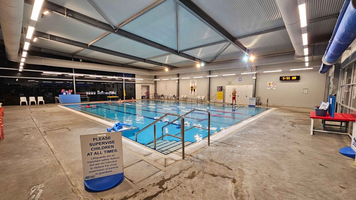 Indoor swimming pool with clear blue water, surrounded by a concrete floor. On the left, there are white plastic chairs and a blue sign reminding to supervise children. In the middle, a metal handrail leads into the pool. On the right, a lifeguard stands near a digital clock showing 5:51 AM. The ceiling has exposed beams and fluorescent lights. A red table with pool equipment is also visible on the right. Indoor swimming pool with clear blue water, surrounded by a concrete floor. On the left, there are white plastic chairs and a blue sign reminding to supervise children. In the middle, a metal handrail leads into the pool. On the right, a lifeguard stands near a digital clock showing 5:51 AM. The ceiling has exposed beams and fluorescent lights. A red table with pool equipment is also visible on the right.