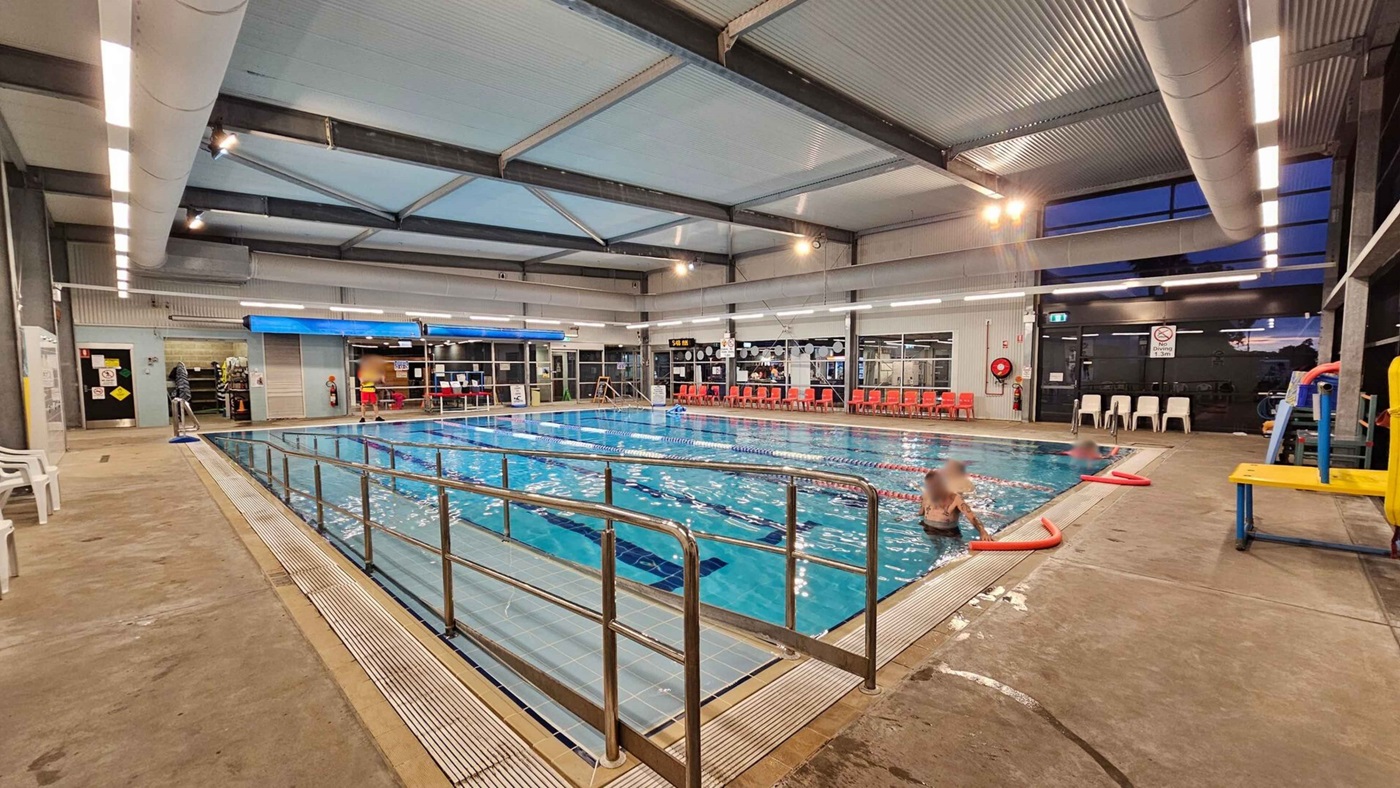 Indoor swimming pool with clear water, surrounded by a metal railing. On the left, there are white plastic chairs and a concrete floor. The right side has a few people in the pool and a yellow platform. The background features a wall with large windows, red chairs, and a lifeguard station. The ceiling is high with exposed beams and lighting fixtures. Indoor swimming pool with clear water, surrounded by a metal railing. On the left, there are white plastic chairs and a concrete floor. The right side has a few people in the pool and a yellow platform. The background features a wall with large windows, red chairs, and a lifeguard station. The ceiling is high with exposed beams and lighting fixtures.