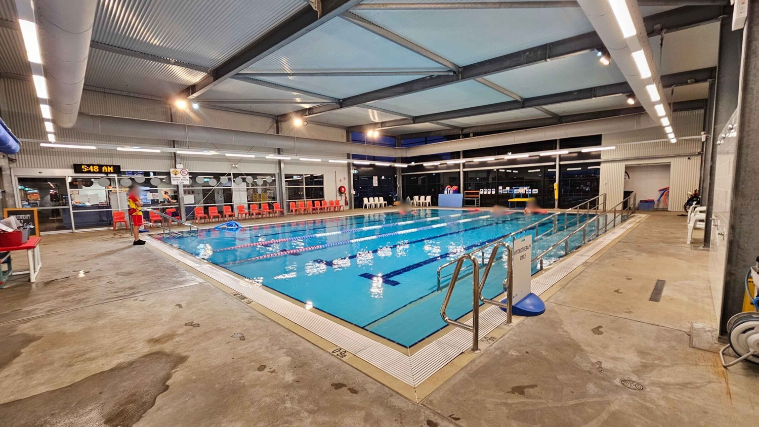 Indoor swimming pool with clear blue water, divided into lanes by floating ropes. On the left, a lifeguard in red and yellow stands by the poolside. Behind them, a digital clock displays 5:48 AM above a row of red chairs. On the right, a metal handrail leads into the pool, with a sign indicating "Hydrotherapy Only." The floor is wet concrete, and the ceiling has exposed beams and lighting. Indoor swimming pool with clear blue water, divided into lanes by floating ropes. On the left, a lifeguard in red and yellow stands by the poolside. Behind them, a digital clock displays 5:48 AM above a row of red chairs. On the right, a metal handrail leads into the pool, with a sign indicating "Hydrotherapy Only." The floor is wet concrete, and the ceiling has exposed beams and lighting.