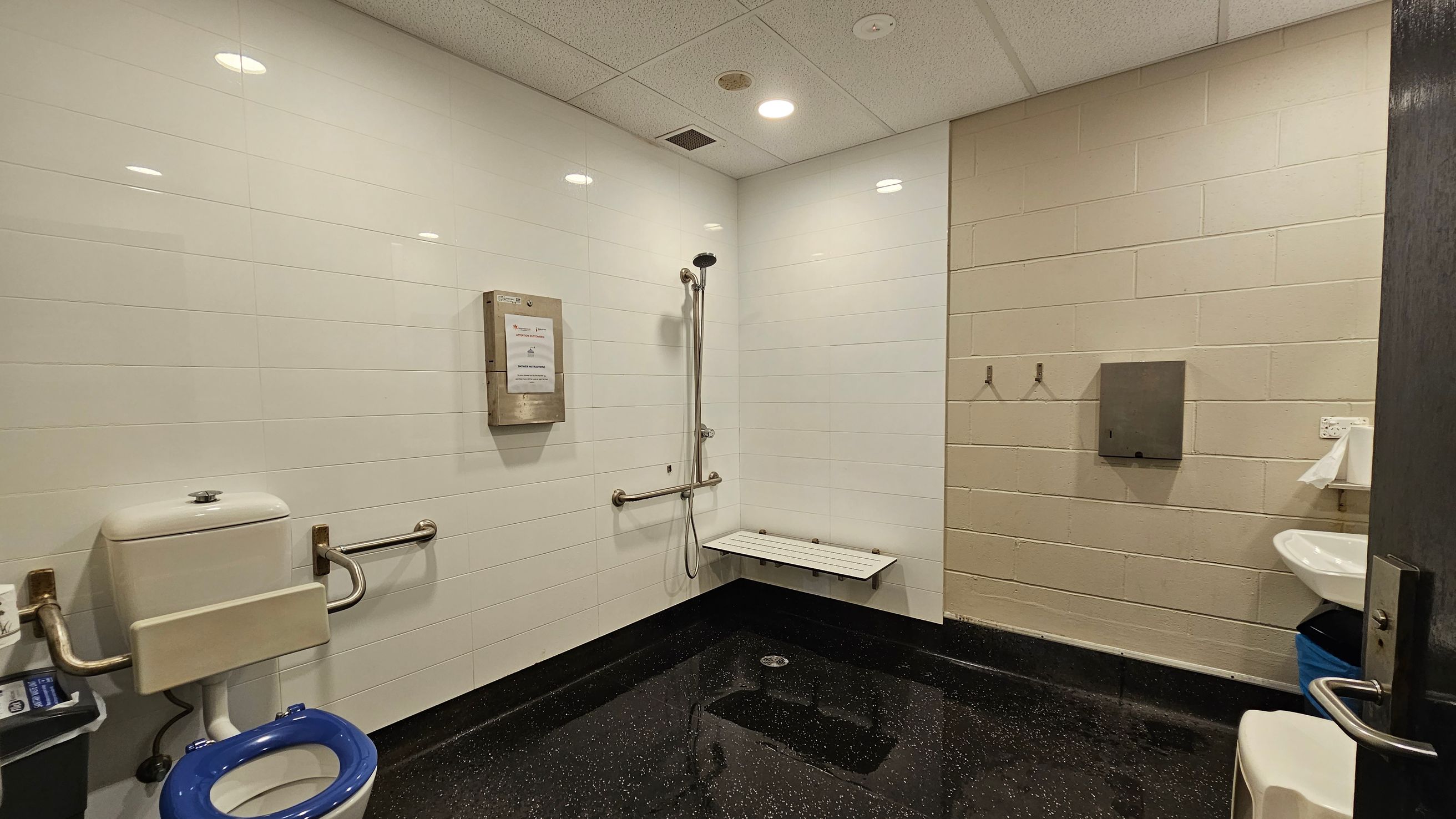 A bathroom with white tiled walls and a speckled black floor. On the left, there's a toilet with a blue seat and support rails. In the centre, a wall-mounted shower with a foldable seat and grab bars. On the right, a sink with a paper towel dispenser above it. Two hooks are mounted on the wall, and a metal box is attached to the wall near the shower. The ceiling is fitted with recessed lights and a vent. A bathroom with white tiled walls and a speckled black floor. On the left, there's a toilet with a blue seat and support rails. In the centre, a wall-mounted shower with a foldable seat and grab bars. On the right, a sink with a paper towel dispenser above it. Two hooks are mounted on the wall, and a metal box is attached to the wall near the shower. The ceiling is fitted with recessed lights and a vent.
