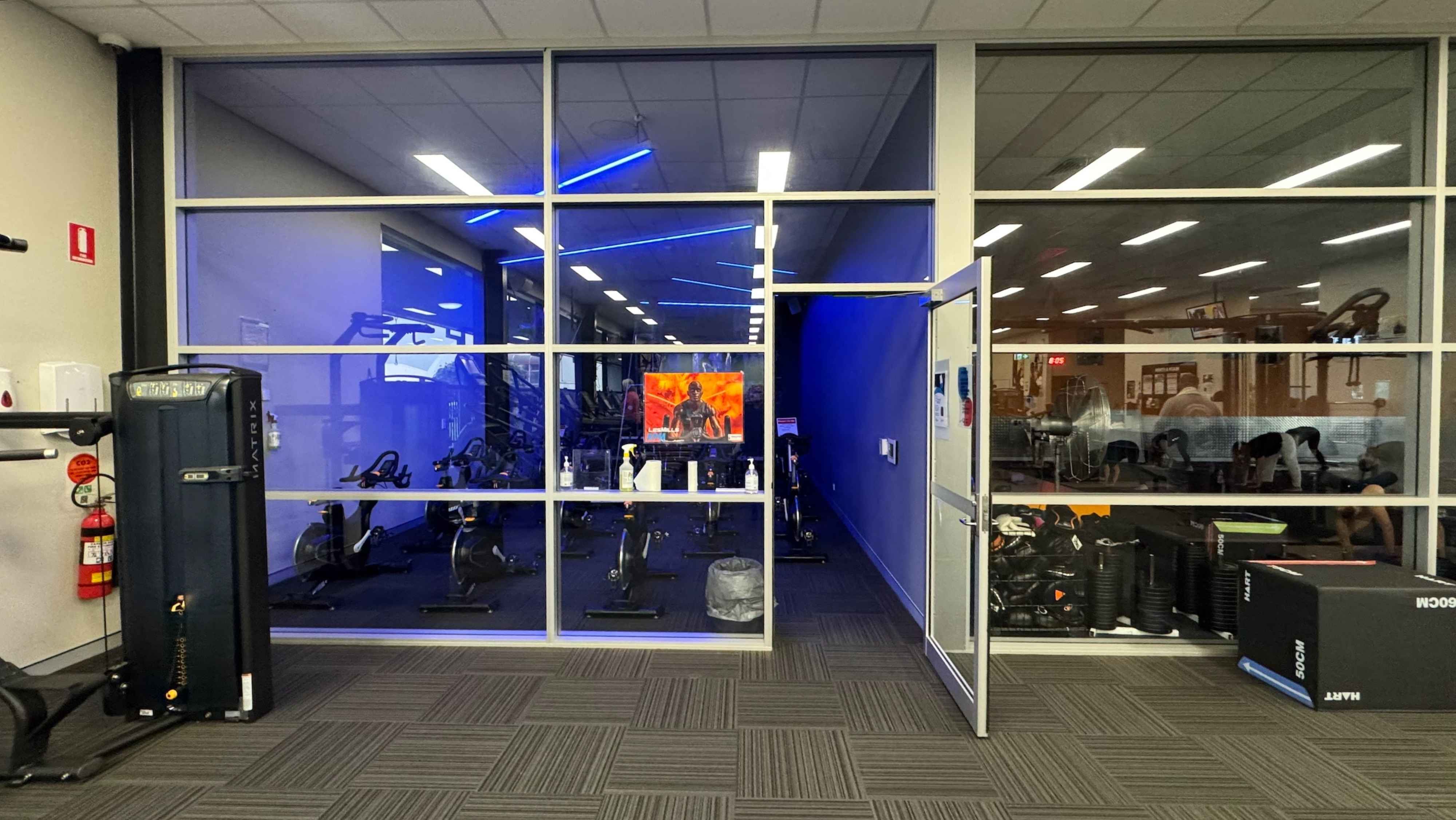 A gym room with a glass wall and door. On the left, there's a black exercise machine and a fire extinguisher. In the middle, through the glass, several stationary bikes are visible, with a TV screen displaying an image of a person. The room has blue lighting. On the right, the door is open, and there are more gym equipment and a large fan. The floor is covered with green patterned carpet tiles, and the ceiling has white panels with fluorescent lights. A gym room with a glass wall and door. On the left, there's a black exercise machine and a fire extinguisher. In the middle, through the glass, several stationary bikes are visible, with a TV screen displaying an image of a person. The room has blue lighting. On the right, the door is open, and there are more gym equipment and a large fan. The floor is covered with green patterned carpet tiles, and the ceiling has white panels with fluorescent lights.