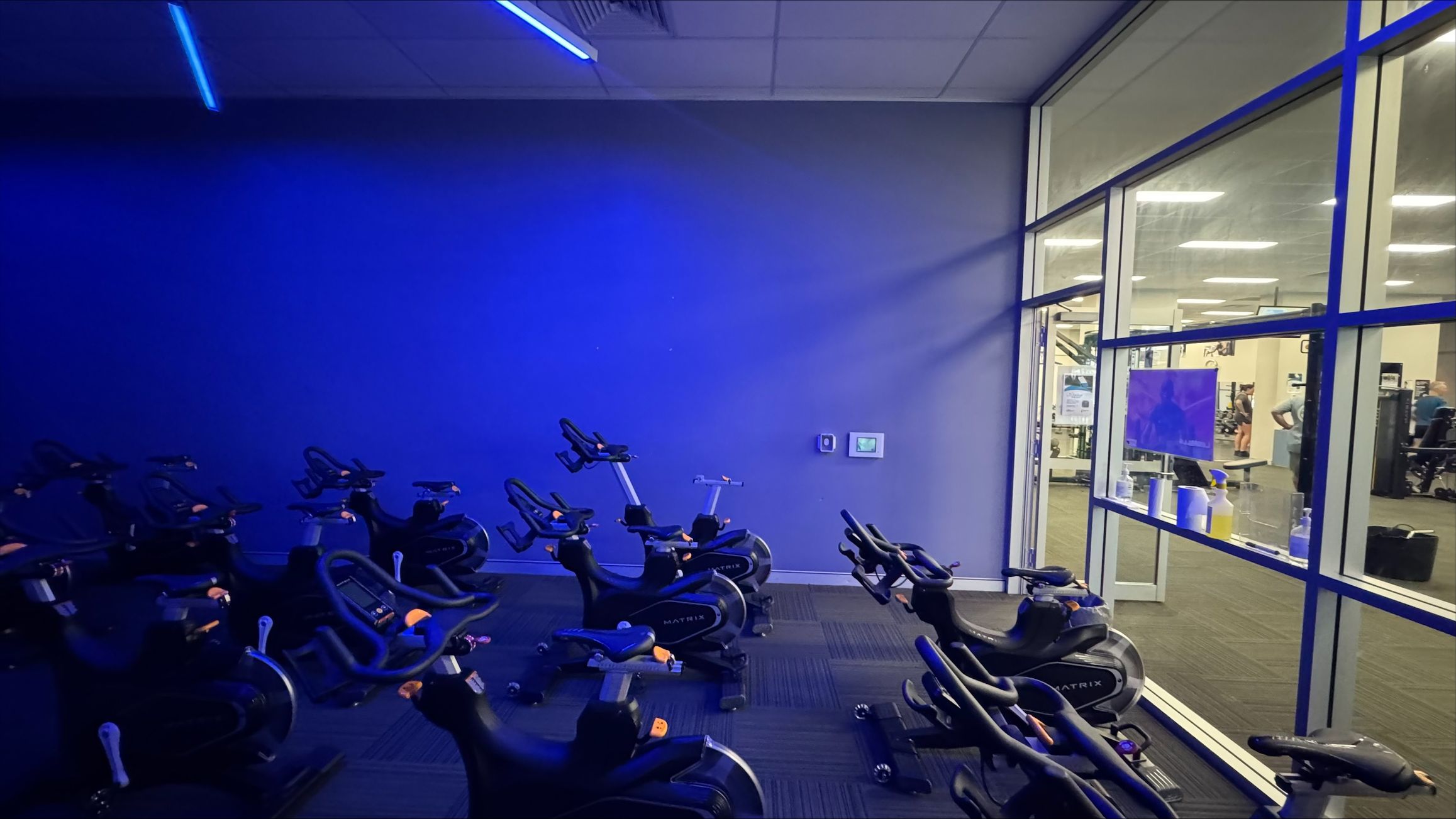 A dimly lit indoor cycling room with several stationary bikes arranged in rows on a dark carpeted floor. The left side of the room is illuminated with blue lighting, casting a glow on the bikes. On the right, a large glass wall separates the cycling area from a brightly lit gym space, where exercise equipment and people are visible. The ceiling has recessed lighting and a blue light strip. A dimly lit indoor cycling room with several stationary bikes arranged in rows on a dark carpeted floor. The left side of the room is illuminated with blue lighting, casting a glow on the bikes. On the right, a large glass wall separates the cycling area from a brightly lit gym space, where exercise equipment and people are visible. The ceiling has recessed lighting and a blue light strip.